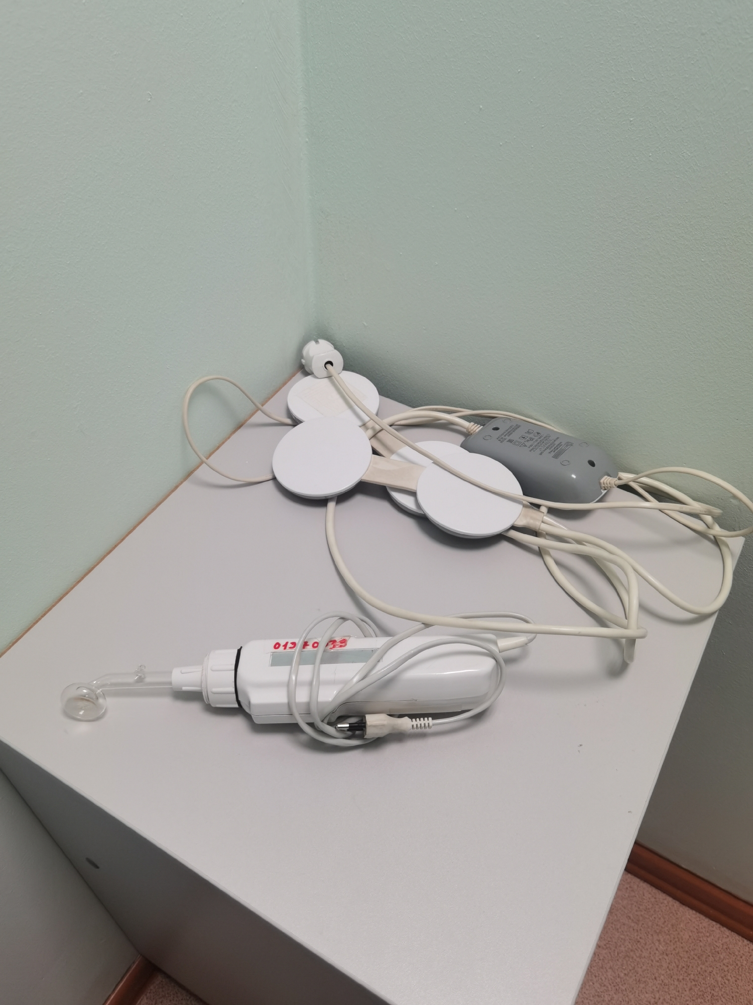 The image shows medical devices on a gray table in a room with light green walls. The devices consist of round, white pads connected by wires to a gray control box, and a white hand-held device with a clear glass attachment. The hand-held device has the inscription "013 f0 39" on its side. The devices are sitting on the table with their wires tangled. No people are visible in the image. The location of the image in the city of Perm, Russia is not deducible from the image.