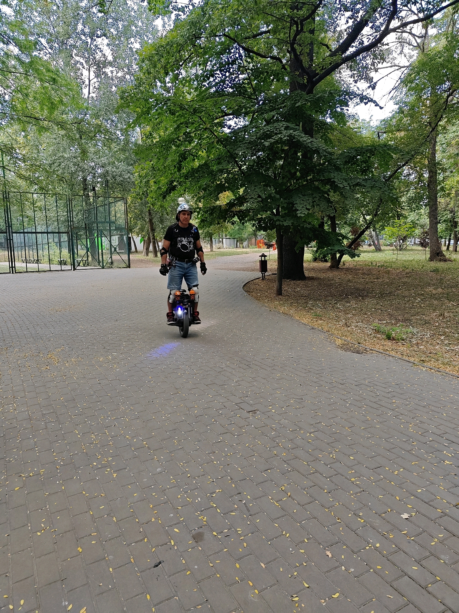 In Rostov-on-Don, Russia, a man rides a unicycle on a paved park path. The man is wearing a helmet, protective pads on his elbows and knees, a t-shirt, and shorts. The unicycle has blue lights on the front. The path is made of gray rectangular pavers and is partially covered with fallen yellow leaves. To the left of the path is a green metal fence and beyond that, more trees. To the right of the path, grass and trees are visible. The sky is overcast.