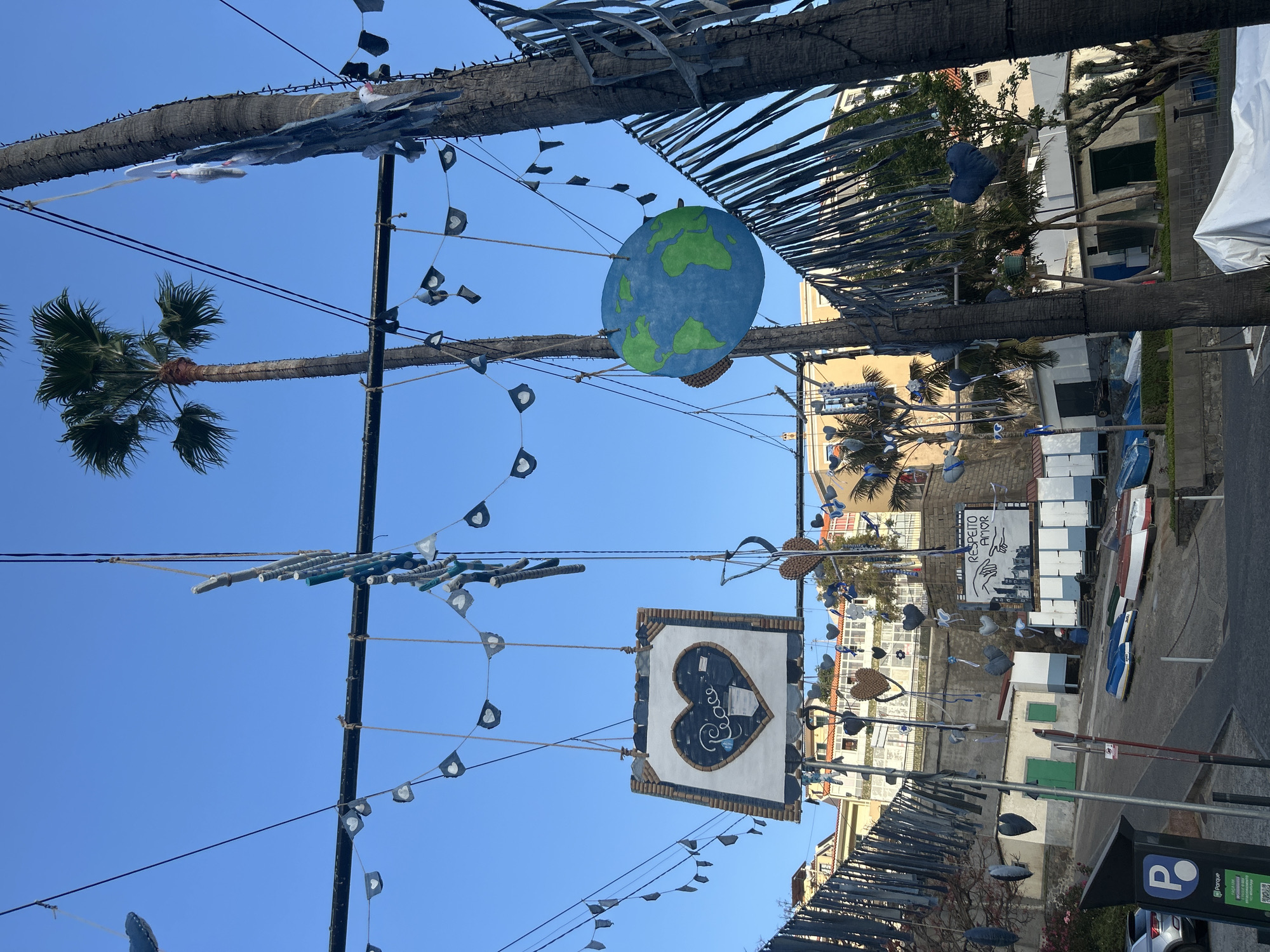 The image captures a street in Câmara de Lobos, Portugal, adorned with decorations. A large, hand-painted globe hangs suspended from the street. Below it, a square sign featuring a heart with the word "Peace" inside is displayed. Decorations including white-and-black pennant flags are hung on cables, which run along both sides of the street. Palm trees are present on the sides of the street, which appear to be decorated with blue-and-white fabric strips. In the background, a sign with the words "RESPEITO AMOR" and hand drawings is visible, along with various white boxes and blue boats. Parked cars are also seen at the bottom right corner of the image. The sky is clear and blue.