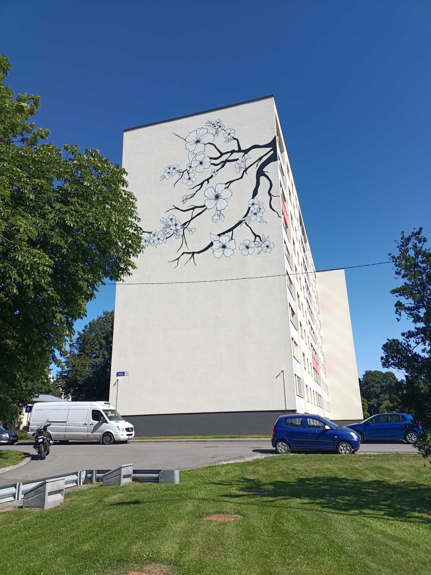 A large apartment building in Tallinn, Estonia, features a mural of a blooming cherry blossom branch on its upper facade. The building is light-colored, with visible windows and some pink accents on the balconies. In front of the building, a street runs alongside a grassy area. A white van, a motorcycle, and two blue cars are parked on or near the street. The sky is clear and blue, indicating a sunny day. Trees are visible on the left and right sides of the frame. A street sign with the text "Kirsi 6" is mounted on a pole near the building.