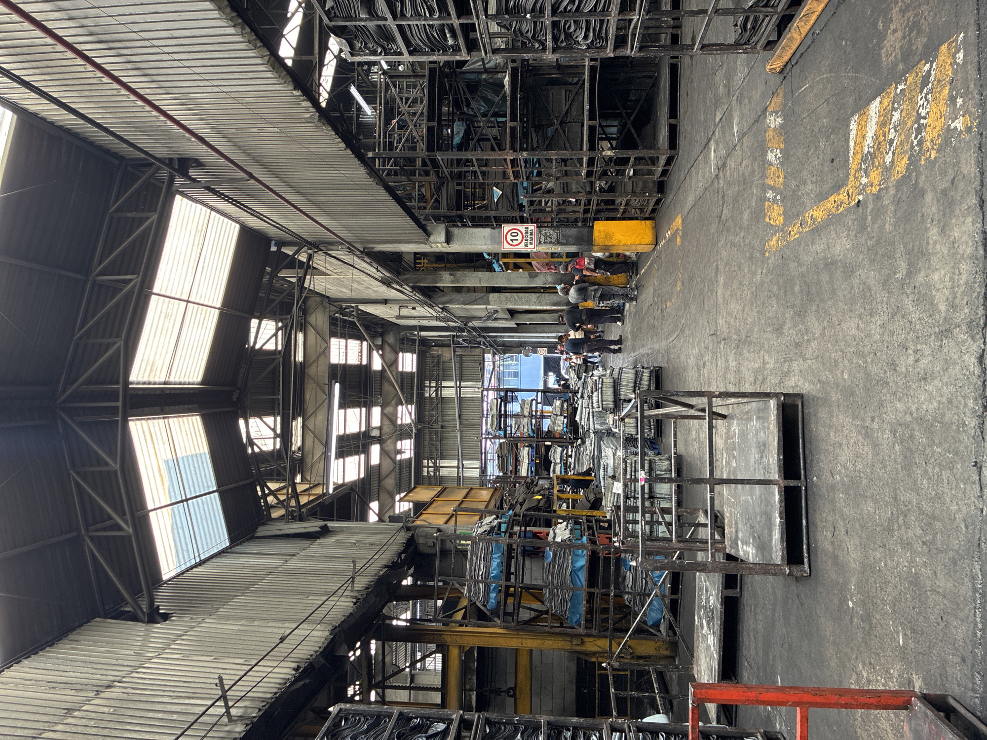 The image depicts the interior of a factory or warehouse in Cuautitlán Izcalli, Mexico. The building has a high, corrugated metal roof with skylights providing natural illumination. The floor is concrete and heavily stained, marked with yellow lines indicating pathways or designated areas. Metal racks and scaffolding dominate the space, stacked high with various items. People are observed in the middle ground, appearing to be workers. The environment suggests a production or storage facility.
