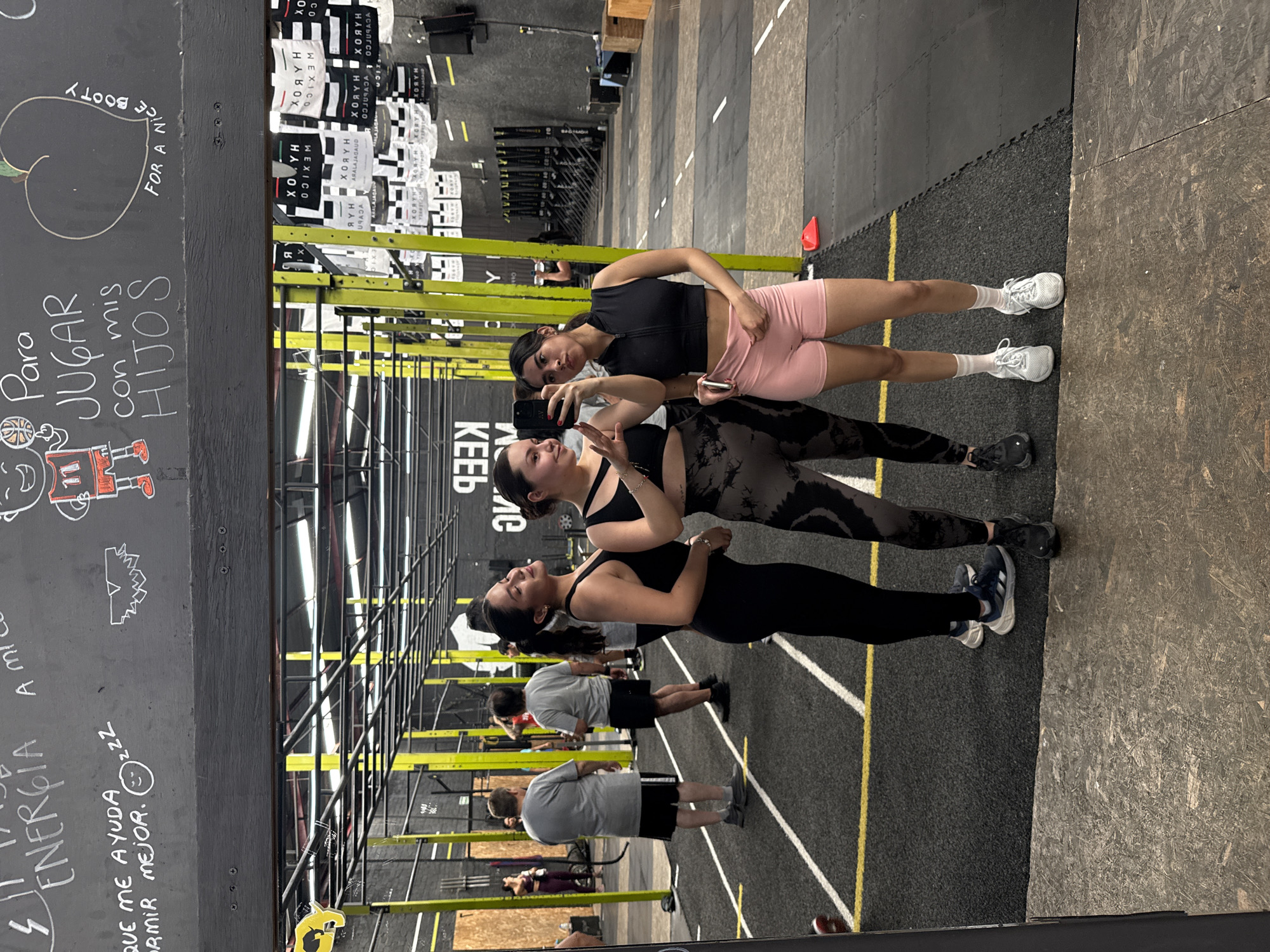 The image depicts a dynamic scene inside a gym, likely a CrossFit facility in Mexico City, Mexico. In the foreground, three women are reflected in a large mirror, with one holding a smartphone to capture a group selfie. The woman with the phone wears a black sports bra and patterned leggings, while the one beside her sports a black top and light pink shorts. The third woman, further back in the reflection, also wears a black top and dark leggings. All three women appear happy and engaged, smiling for the camera.
The gym environment features a dark rubber-matted floor with distinct yellow and white lines, typical of training zones. Various workout equipment, including metal racks and bars, are visible in the background, where several men are also seen, some engaged in exercise.
To the left, a dark chalk wall displays handwritten messages in Spanish, adding local flavor. These include "Para JUGAR con mis HIJOS" (To play with my children) and "QUE ME AYUDA ENERGIA DORMIR MEJOR. ZZZ" (that helps me sleep better energy sleep better ZZZ). There's also an English phrase, "FOR A NICE BOOTY," accompanied by a drawing of a peach. In the upper background, stacked items or banners prominently display text such as "XO HABOX WEXICO," "XOYH," "ARALAJAGAUD," and "USADA," clearly indicating the Mexican location. The scene captures a moment of active social interaction within a fitness setting.