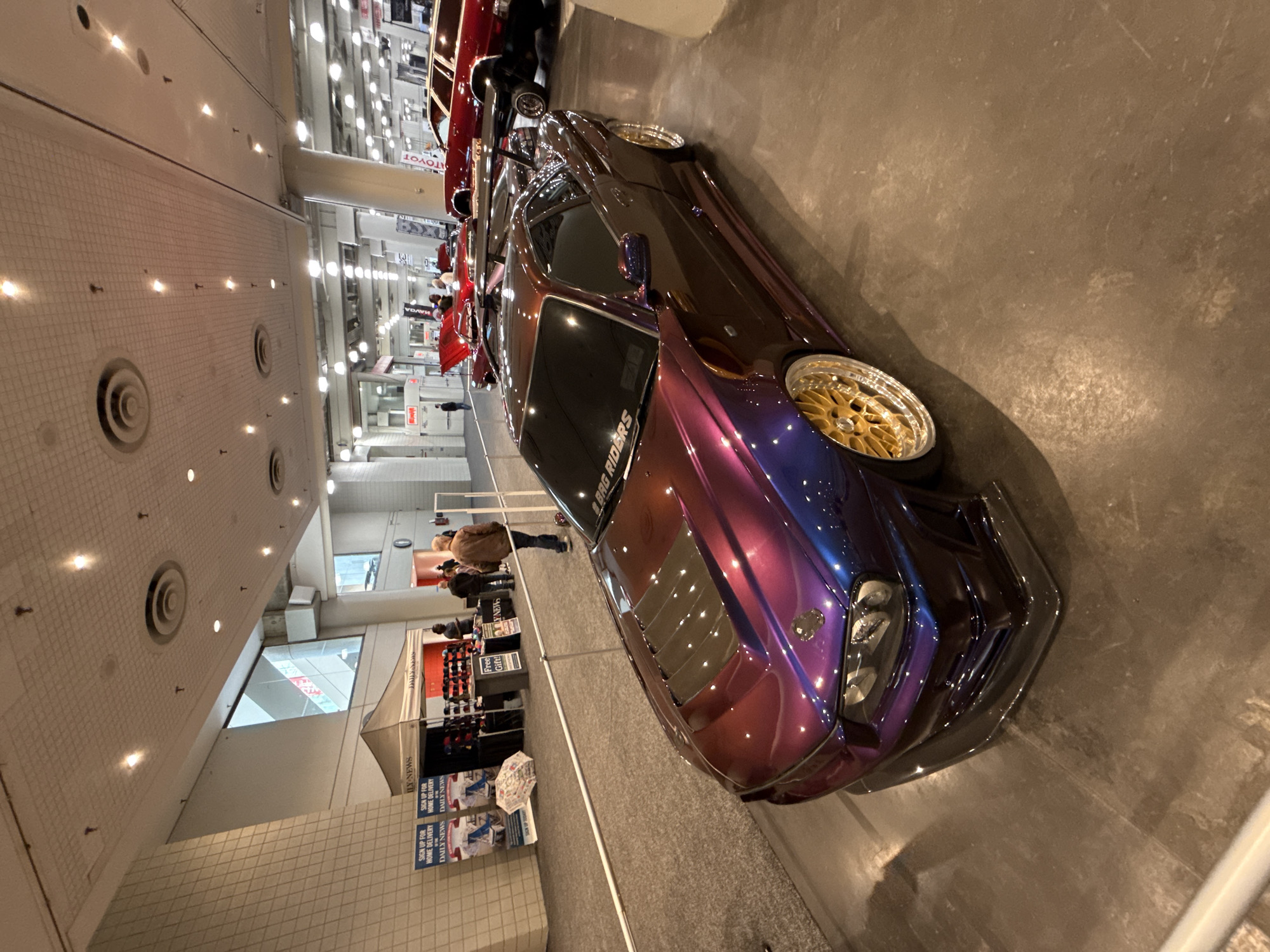 The image captures a car show or automotive exhibition at a convention center in New York City, United States of America. Dominating the foreground is a customized car with a unique color-shifting paint job, possibly purple, blue, and maroon. The car features gold rims and an aftermarket front bumper.
In the background, multiple booths are set up, some displaying merchandise for sale. People are browsing these stalls and walking around the exhibit. A sign for "ILY NEWS" and "BAG RIDERS" is visible. The convention center has a tiled floor and a high ceiling with circular air vents and recessed lighting.