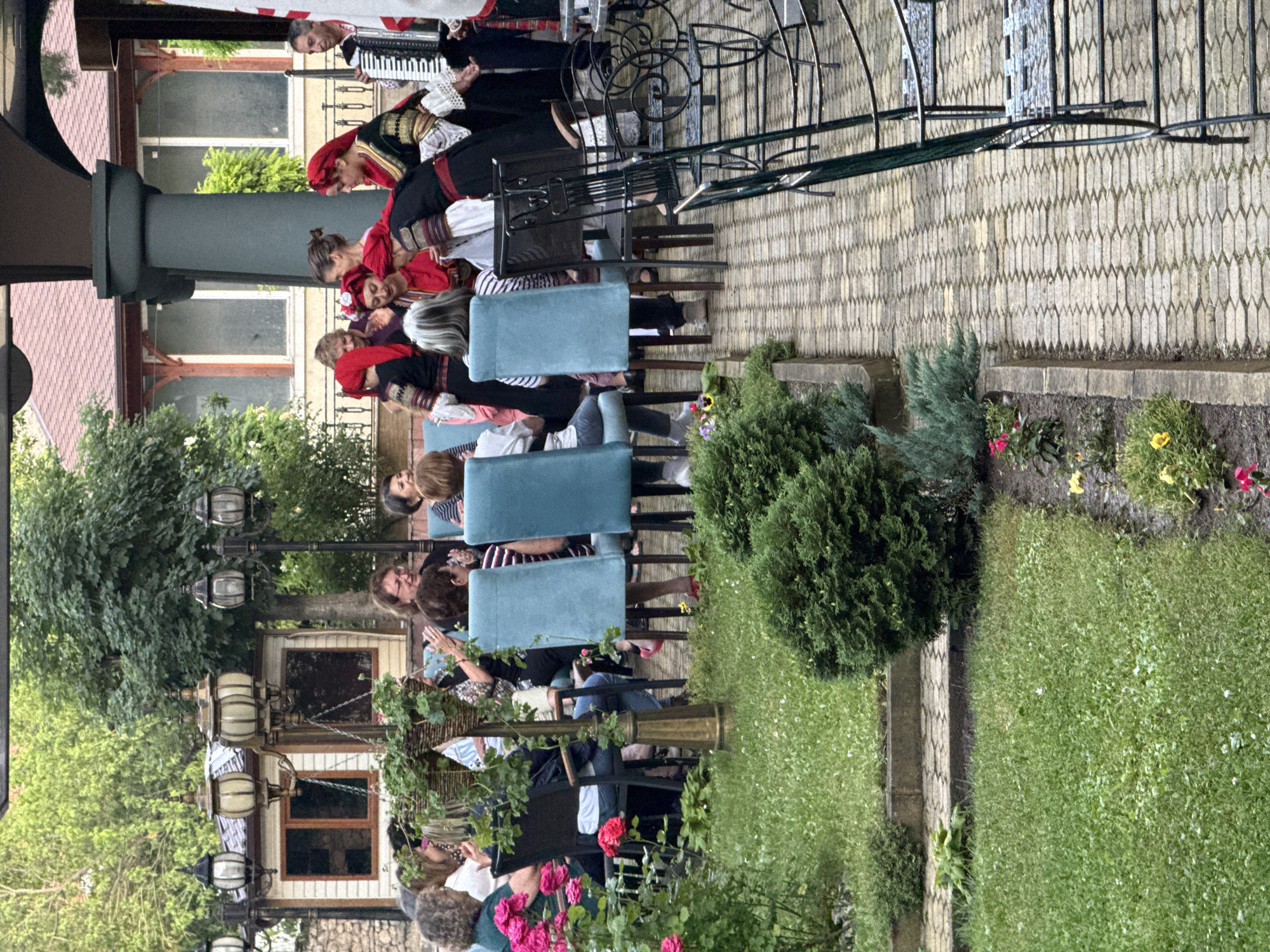 A group of people are gathered at an outdoor restaurant in Kyustendil, Bulgaria. Several women are seated at tables, some of whom are clapping. A group of people in traditional Bulgarian folk costumes are performing, including a man playing an accordion. The setting appears to be a courtyard with a paved walkway, grassy areas, and lush greenery. There are outdoor light fixtures and furniture, suggesting a pleasant evening gathering.