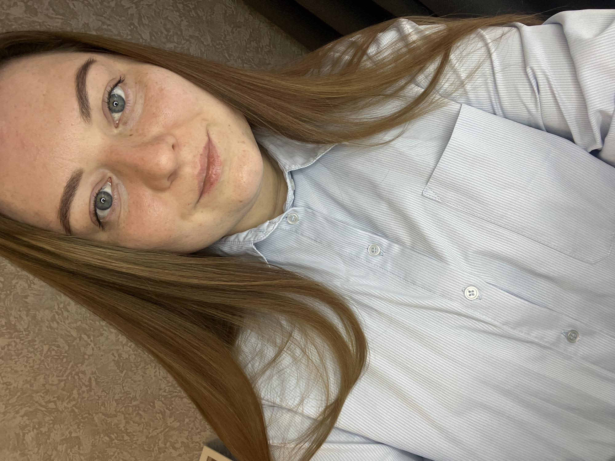 A woman with long, light brown hair is lying on her side, wearing a light blue and white striped button-down shirt. She has fair skin, freckles, and light-colored eyes. The background includes a textured wall and a dark-colored surface. There is no indication of the location of Раздолье, Russia.