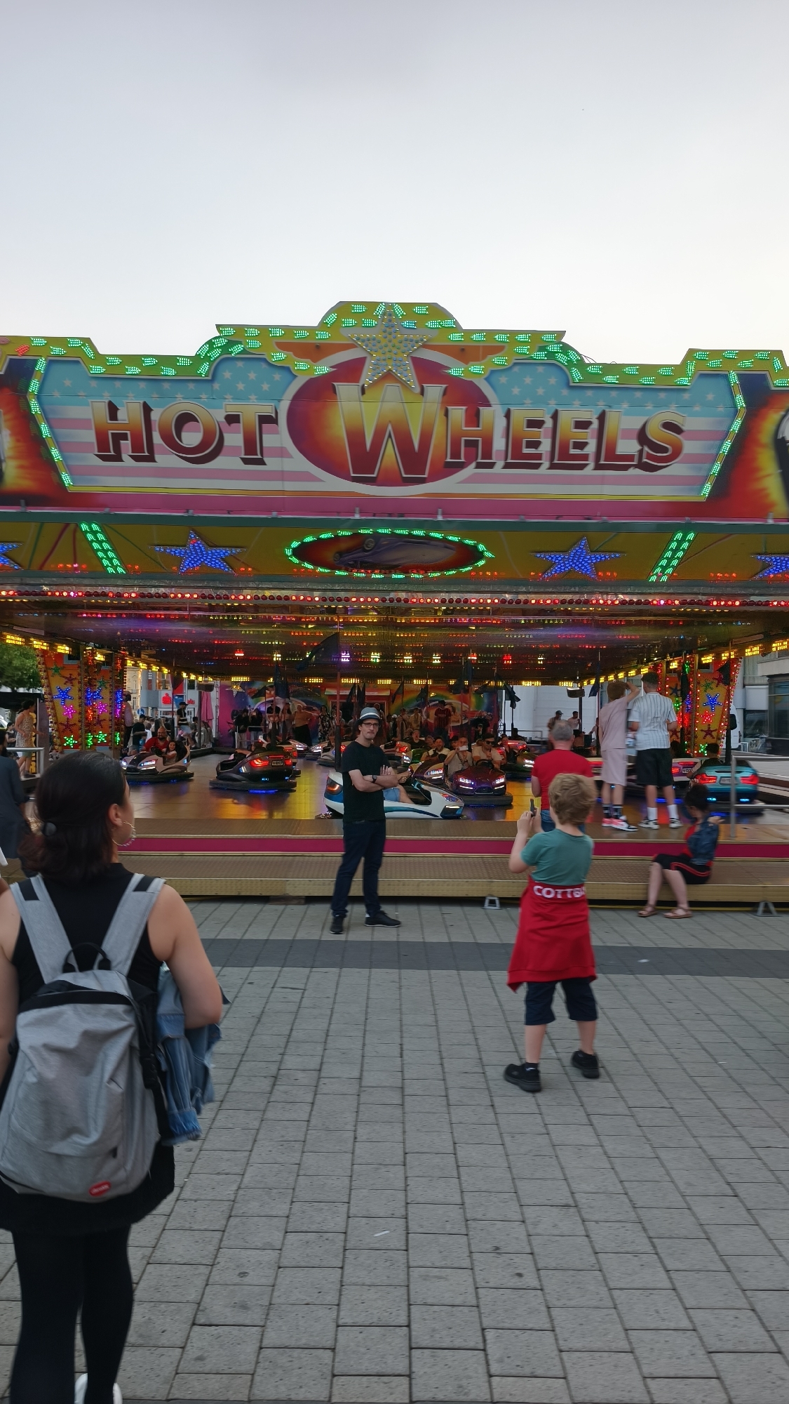 The image shows a bumper car ride at an outdoor amusement area. A large sign at the top of the ride reads "HOT WHEELS" with colorful lights. Inside the ride area, several bumper cars are in motion, and various people are visible riding in them or standing around the platform. In the foreground, a woman wearing a black top and a grey backpack is facing away from the camera, walking to the left. To her right, a young boy wearing a red apron and dark shorts is standing and holding a mobile device, appearing to take a photo. Another man in a dark t-shirt and cap stands with his arms crossed, facing the bumper car ride. The ground is paved with grey interlocking bricks. The lighting suggests it is late afternoon or early evening.