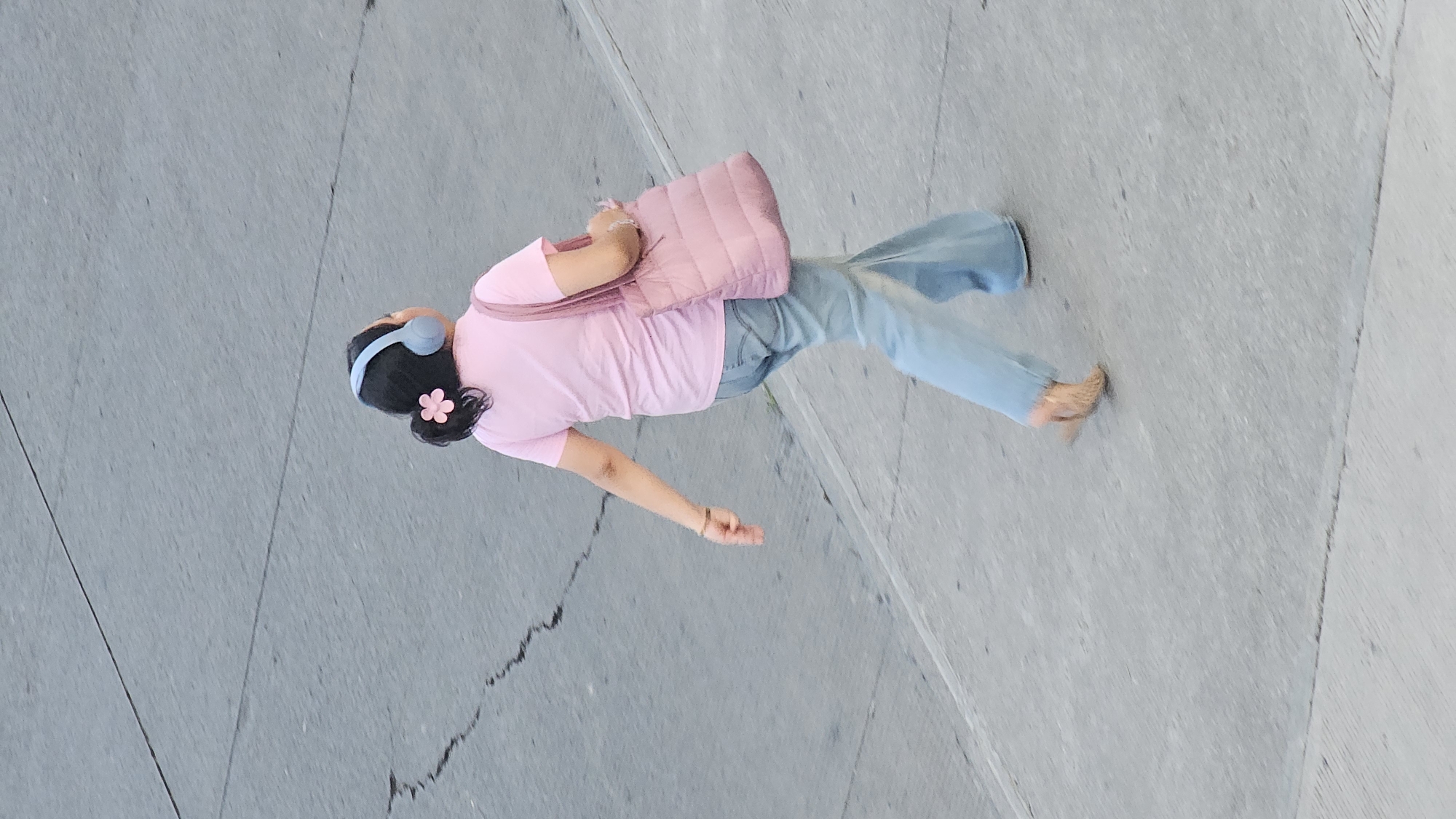 From a high-angle perspective, a woman is walking on a concrete surface. She is wearing light blue headphones and has a pink flower in her dark hair, a pink t-shirt, light blue jeans, and tan high heels. A light pink tote bag is slung over her shoulder. The concrete surface has visible seams and slight variations in shade. The location is likely Puebla, Mexico.