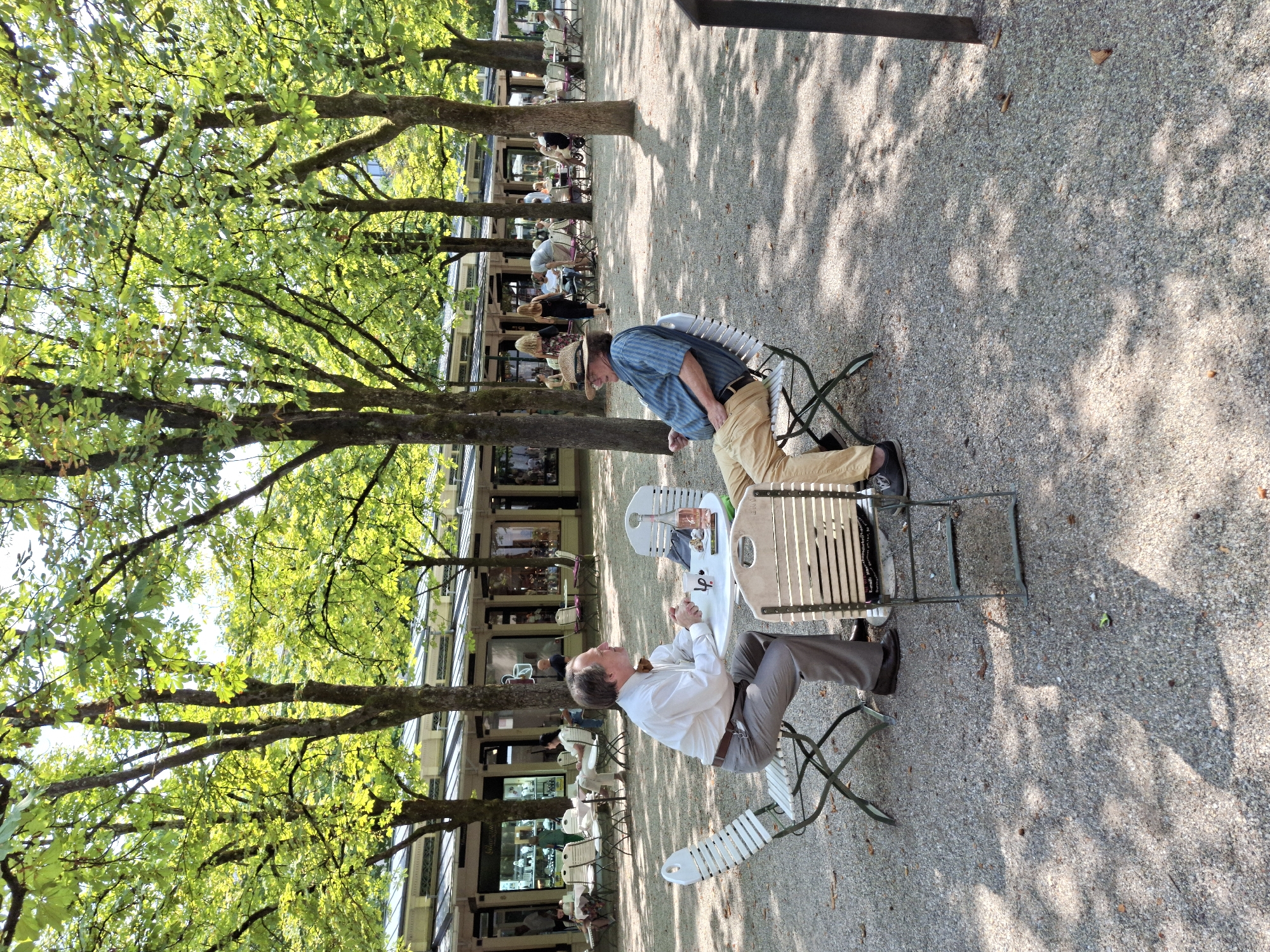 In Baden-Baden, Germany, two men are seated at a small round table in an outdoor seating area with a gravel ground. The man on the left, wearing a white collared shirt and grey pants, is looking to the right and appears to be speaking. The man on the right, wearing a blue and white striped shirt, khaki pants, and a straw hat, is looking towards the man on the left and has his hand raised slightly. On the table between them are two cups, a bottle of rosé wine, and what appears to be a small bag.
Behind them, a line of shops with awnings stretches across the background, and trees with lush green leaves provide shade, creating dappled sunlight on the ground. Other people are visible in the distance, seated at tables and walking. The scene suggests a relaxed atmosphere in a public space, likely a cafe or restaurant with outdoor seating.