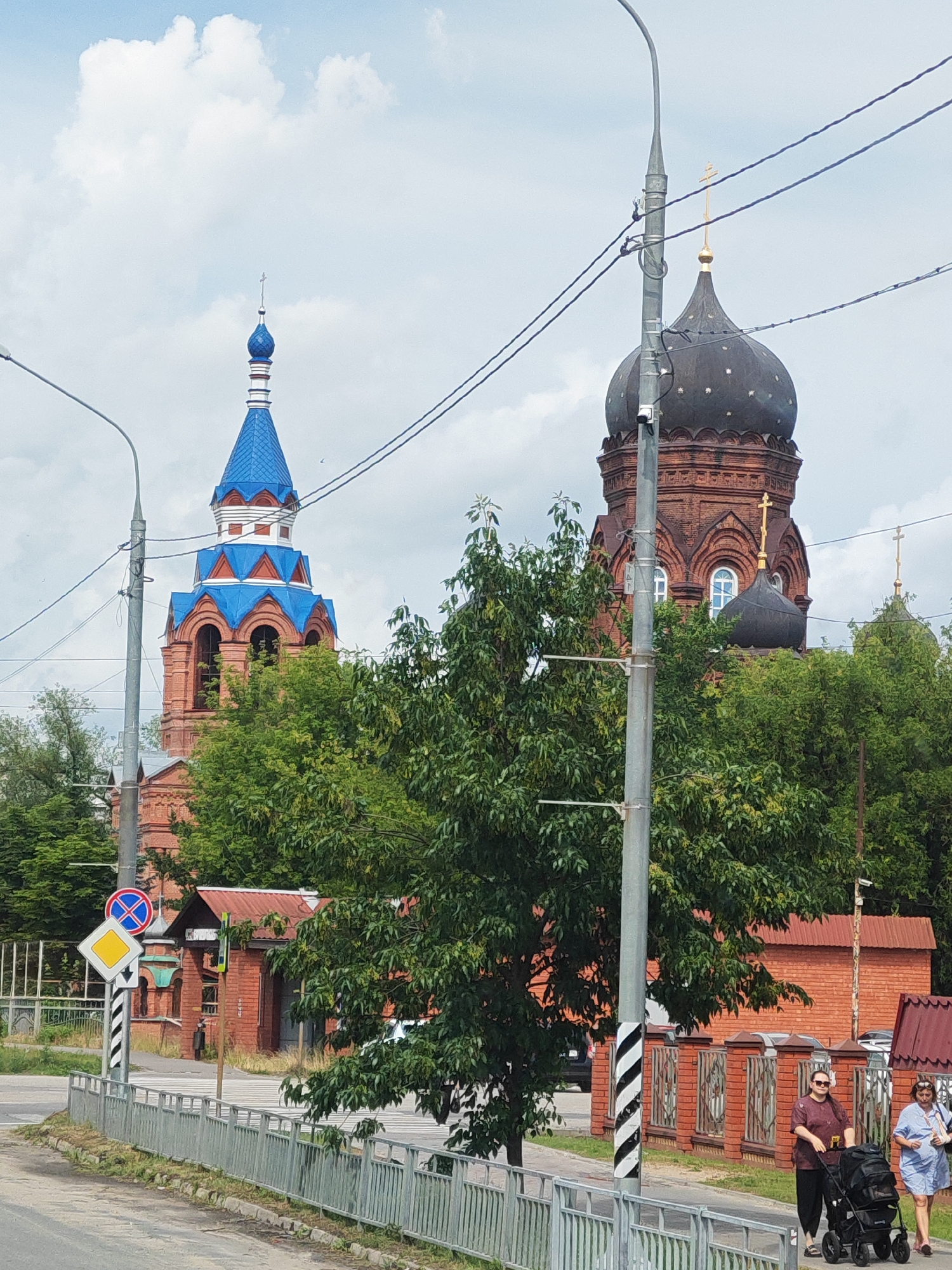 In Likino-Dulyovo, Russia, a street scene unfolds with two churches prominently featured. The larger church, with its dark dome and brick facade, is partially obscured by lush green trees. To its left, a smaller, white-and-blue domed church tower rises, also surrounded by foliage. A metal fence runs along the foreground, bordering a sidewalk where two women are visible. One woman, wearing dark clothing and sunglasses, pushes a black stroller. The other woman, in a light blue dress, walks beside her. A traffic light pole with diagonal black and white stripes stands near the women. In the background, a red-brick building and another smaller church structure can be seen. The sky above is partly cloudy.