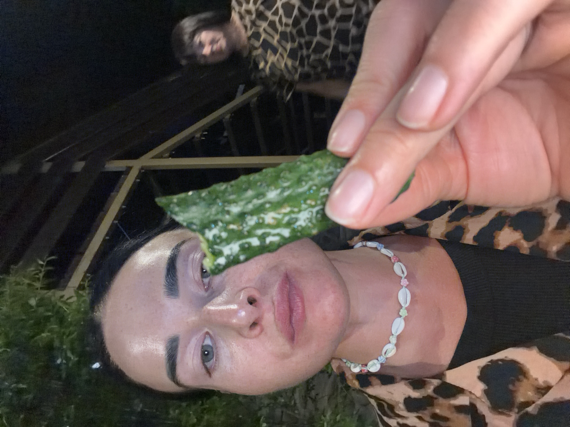 A close-up, point-of-view shot shows a woman holding a piece of green, textured food with white speckles up to her face. Her face is partially obscured by the food item. She is wearing a leopard-print shirt and a seashell necklace. In the background, another woman with dark hair is visible, wearing a patterned top. The setting appears to be outdoors at night, possibly a restaurant or cafe, with foliage and wooden structures visible. There is no identifiable information to indicate the location is Sochi, Russia.