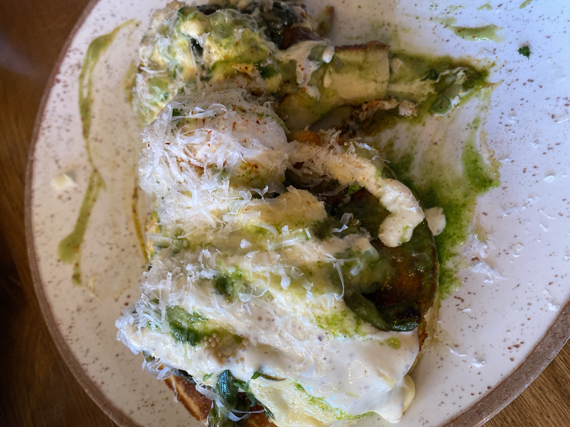 The image is a close-up shot of a plate of food. Two stacks of toasted bread appear to be covered in white sauce and shaved cheese. Green sauce is drizzled on top. The plate is cream-colored with brown specks and a brown rim. The food appears to be partially eaten. The background is blurry. There is no indication of Strasbourg or people present in the image.