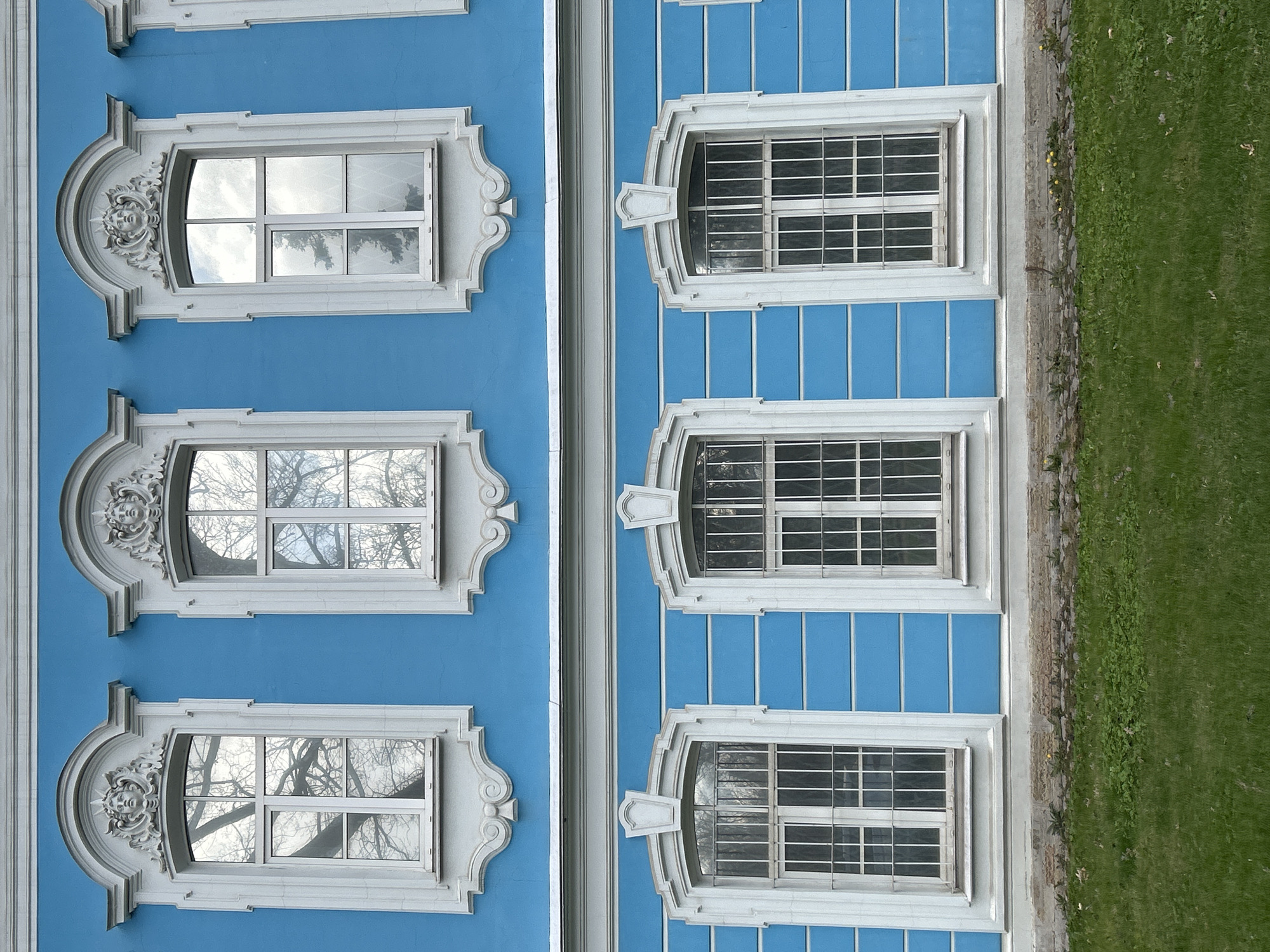 The image shows a section of a building in Saint Petersburg, Russia, likely a palace or significant historical structure, characterized by ornate window frames and a vibrant blue facade. The building is distinguished by two different window styles. One style is highly decorative, featuring baroque-style elements and ornate sculpted faces above each window. These windows reflect the sky and bare tree branches. The second window style, to the right, is simpler and more geometric, with a white frame and multiple panes, some windows have an additional layer of bars. The building's facade is painted in a bright blue hue with vertical paneling. A narrow strip of greenery, likely grass, runs along the edge of the building. The overall impression is of a well-maintained historical structure, capturing the architectural richness of Saint Petersburg.