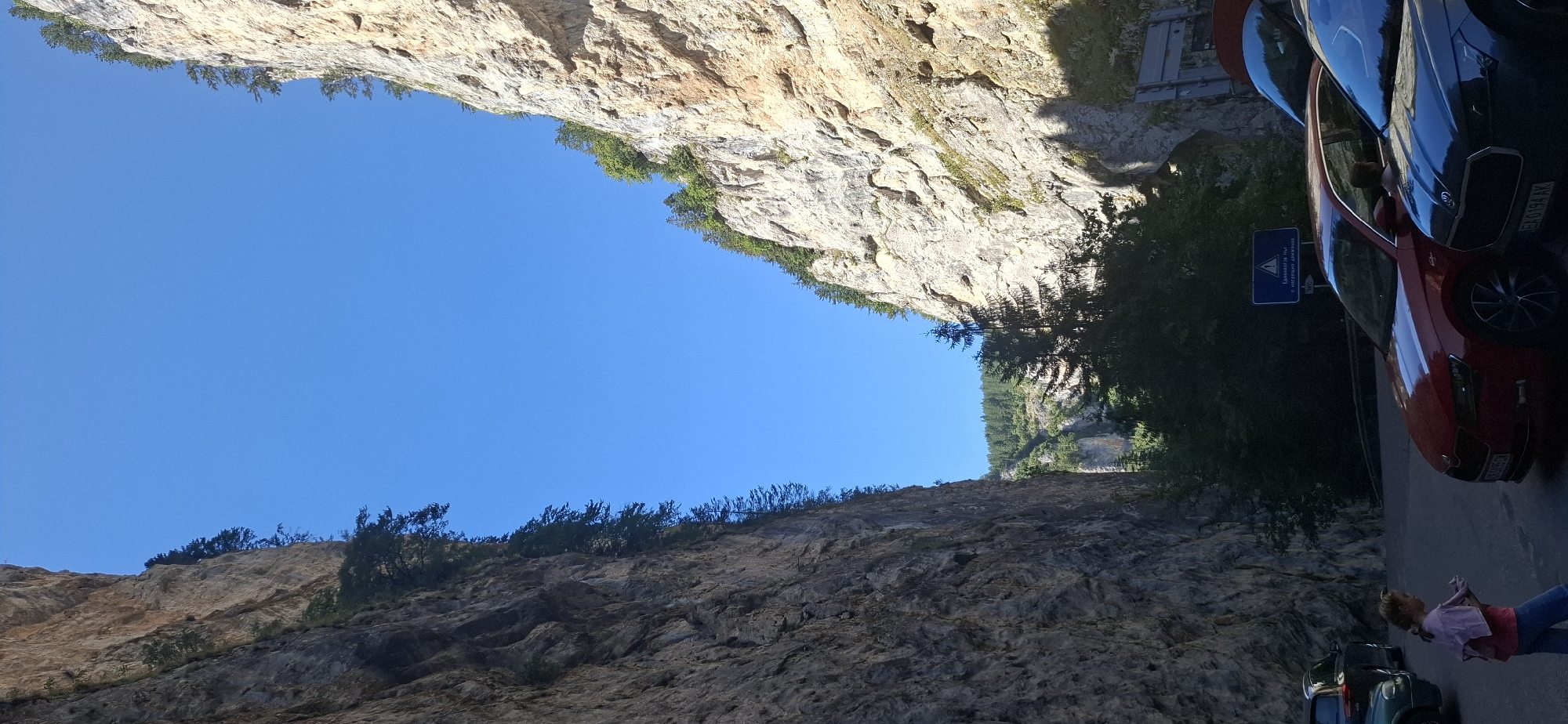 The image shows a narrow canyon with steep, rocky cliffs on either side, under a clear blue sky. A paved road runs through the canyon, with several cars parked on it. In the foreground, a young girl wearing a pink shirt and blue jeans stands near a red car. A sign with Bulgarian text and a WC symbol is visible on the right side of the road, near a concrete structure. The overall impression is of a scenic natural location, possibly a tourist destination, with evidence of human presence and infrastructure. The location is the Trigrad Gorge in Bulgaria, a popular natural attraction known for its dramatic rock formations and the nearby Trigrad Caves.