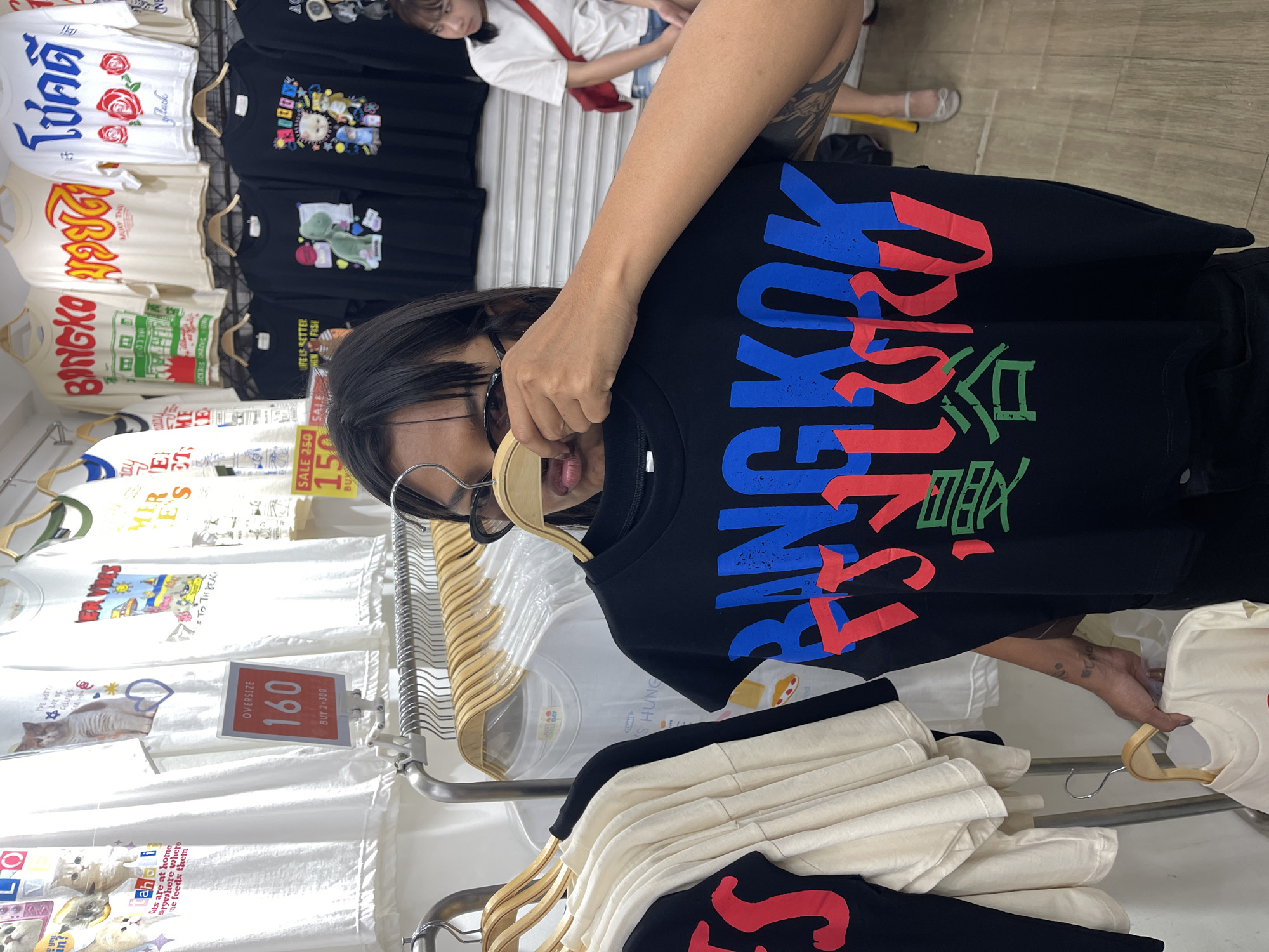 This media file shows a person in a clothing store, likely in Bangkok, Thailand, given the visible text. The person is holding up a black t-shirt with colorful text in Thai and Japanese characters. The t-shirt reads "BANGKOK" in large blue and red letters, followed by what appears to be Thai script and Japanese characters in green. The person is wearing glasses and has dark hair.
The background reveals a display of various t-shirts hanging on a rack. Some of the t-shirts are white with different graphics and text, including one with roses and the word "luck," and another with cartoon characters. Another t-shirt features a graphic of a cat and the text "cats are at home everywhere where the owner feeds them." Price tags are visible, with one indicating "OVERSIZE 160 BUY 2-300."
In the upper background, another person is seen wearing a white t-shirt and denim shorts, with a red strap across their body. The store appears to be indoors, with wooden flooring and a bright, possibly artificial, light source. The overall scene suggests a casual shopping environment. The image is oriented sideways, with the main subject in the foreground.
