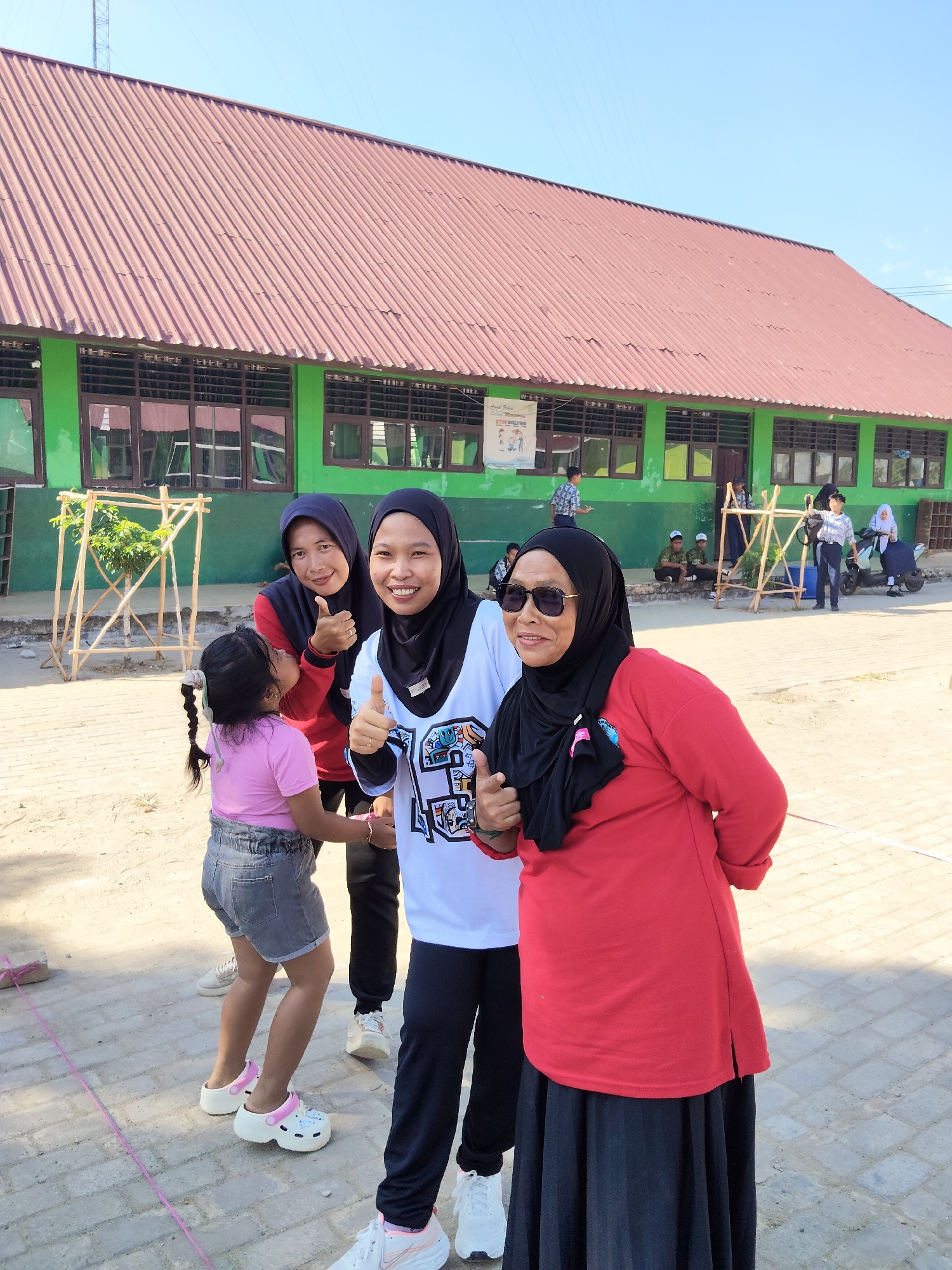 In Lori, Indonesia, a group of individuals, primarily women wearing hijabs, pose for a photo outside a green building with a red corrugated metal roof. The building appears to be a school, with visible windows and a banner on the left wall that reads "Lain Dalam Memantau STOP BULLYING".
In the foreground, three women are prominently featured. The woman on the far right wears a red long-sleeved shirt, a black hijab, and a black skirt, with sunglasses on. She is giving a thumbs-up. Next to her, a woman in a white t-shirt with black sleeves and a black hijab also gives a thumbs-up. To the left of these two, another woman in a red long-sleeved shirt and a blue hijab smiles and gives a thumbs-up.
A young girl in a pink t-shirt and grey denim shorts stands between the women, looking up. Other individuals, including children and adults, are visible in the background, some sitting on the ground and others standing near a wooden structure. The ground is paved with bricks and sand. The scene suggests an outdoor event or gathering.