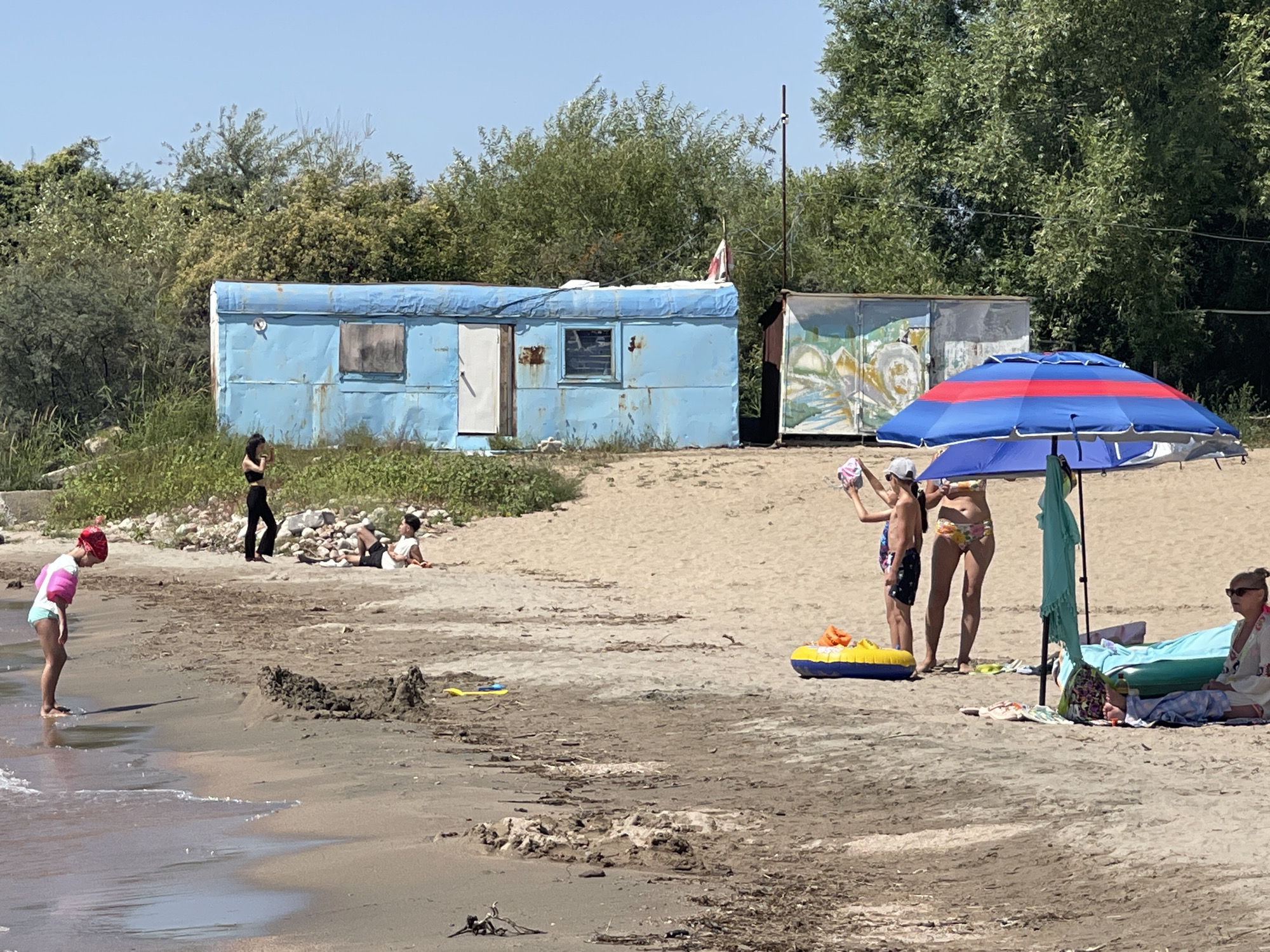 The image depicts a beach scene with two dilapidated structures in the background. On the left, a blue, rectangular portable building with a weathered exterior and visible damage stands against a backdrop of green trees and a clear blue sky. A small, red and white flag is attached to a pole near the top of this structure. To its right, another similar, but smaller, structure is visible, decorated with a colorful mural.
In the foreground, the sandy beach is populated by several individuals. A young child wearing a pink and turquoise swimsuit is standing near the water's edge, looking down at the sand. Further back, two adults are seated on the sand. One adult is lying down, while the other is sitting up and appears to be looking at something. To the right, under a striped blue and red umbrella, a group of people are gathered. One person is holding a pink object, another is standing with a yellow and blue inflatable raft nearby, and a woman is seated on a beach towel, wearing sunglasses. A light turquoise towel is draped over the umbrella's pole. The overall environment is a beach with scattered debris on the sand and vegetation in the background. The location is identified as Ornek, Kyrgyzstan.