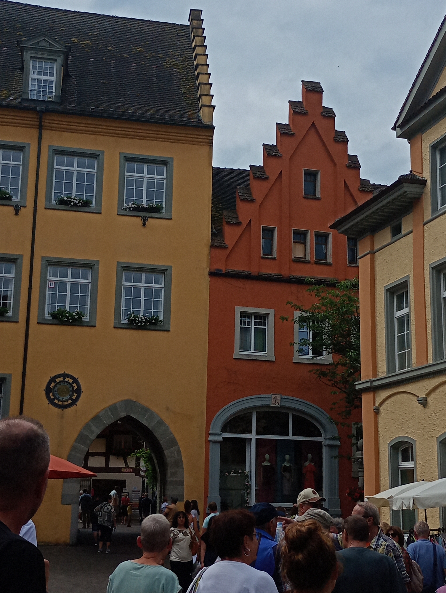In Meersburg, Germany, this image captures a view of a street with historic buildings and pedestrians. The foreground shows the backs of several people, with one person wearing a black t-shirt and another in a light blue shirt. In the middle ground, an arched gateway is visible on a yellow building, with people walking through it into a shaded area. The gateway is adorned with a circular decorative element above it. Adjacent to the yellow building is an orange building with a distinctive stepped gable. Further back, more people are seen browsing at stalls beneath a striped awning. The sky is overcast. The buildings exhibit traditional European architecture with multiple windows, many featuring flower boxes.