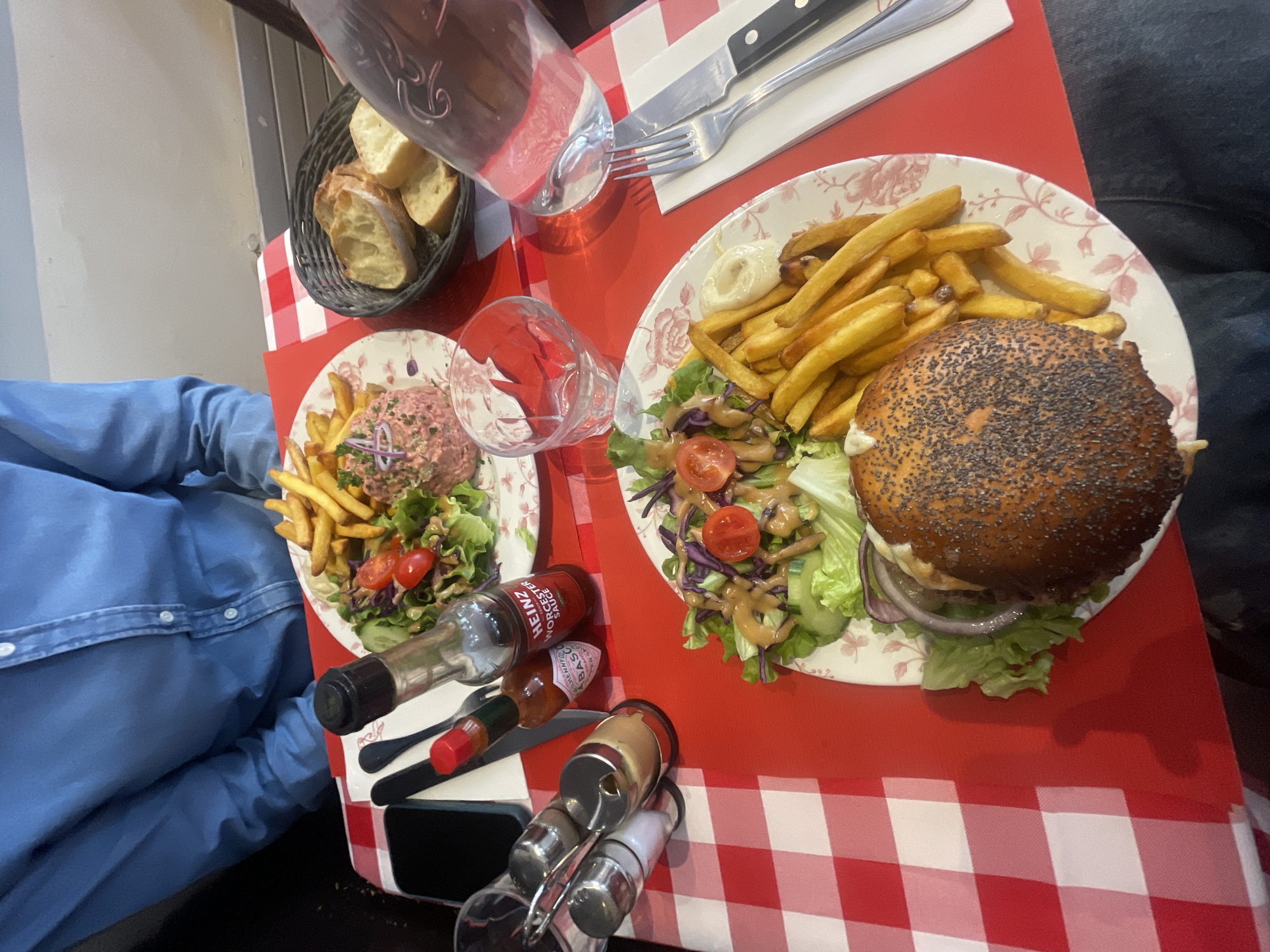 The image captures a meal scene, likely in a restaurant in Paris, France, suggested by the Parisian-style red and white checkered tablecloth. On the table are two floral patterned plates, one with a burger topped with a poppy seed bun, french fries, and a side salad, and the other with what appears to be steak tartare, french fries, and a side salad. A basket of bread, a bottle of water, and drinking glasses are also present. Condiment bottles, including Heinz Worcestershire sauce and Tabasco, are visible along with salt and pepper shakers. Silverware is laid out for dining. A portion of a person's blue shirt sleeve is visible on the left, and another person wearing denim is on the right. The overall setting suggests a casual dining experience.