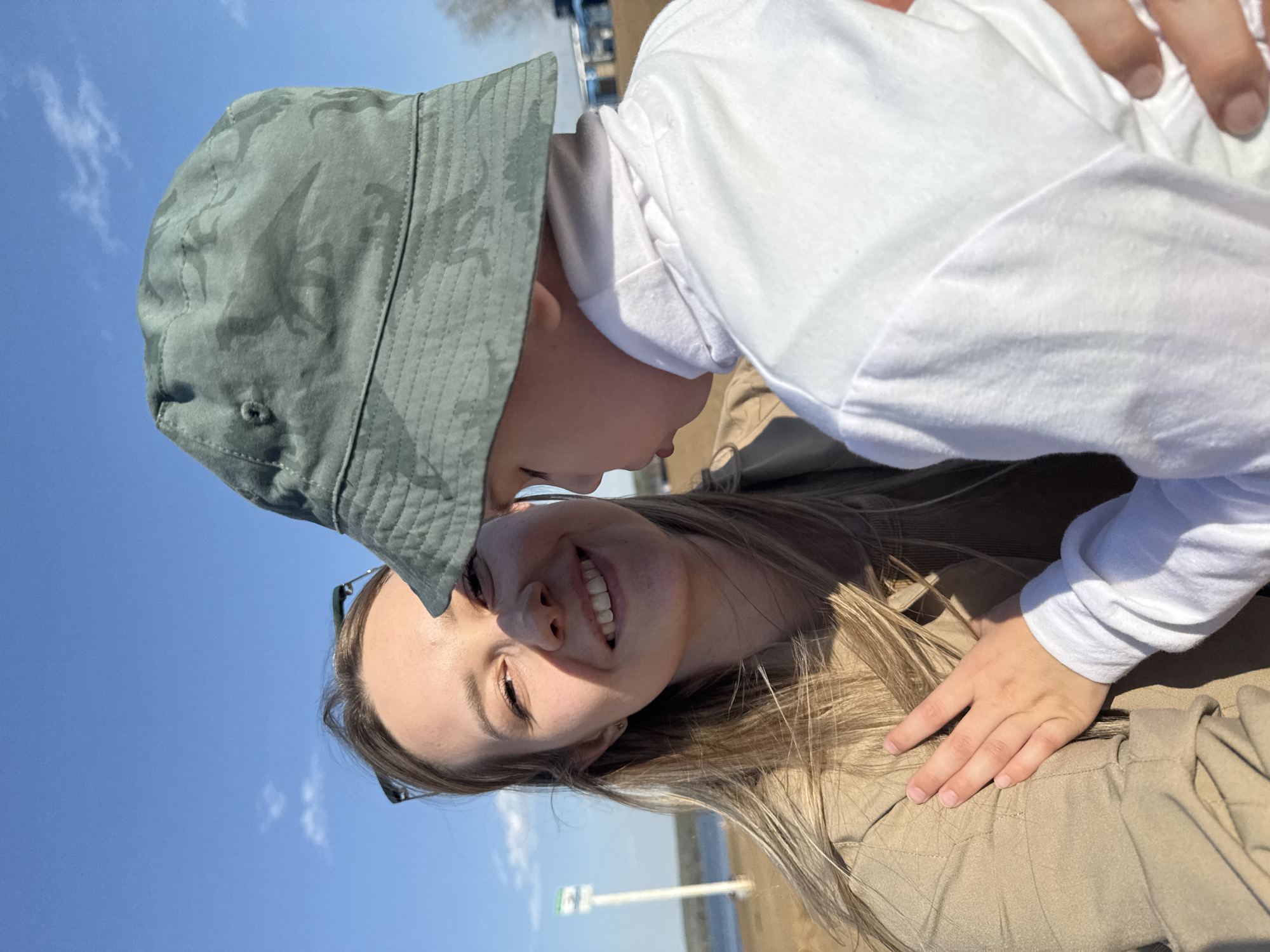 A photograph taken outdoors in Chaikovsky, Russia, shows a woman and a child in close proximity. The woman, with light skin and blonde hair, is smiling. She is wearing sunglasses pushed up on her forehead and a tan jacket. A young child wearing a white long-sleeved shirt and a green patterned bucket hat is leaning towards her. The sky is blue with few clouds. In the background, there is a body of water.