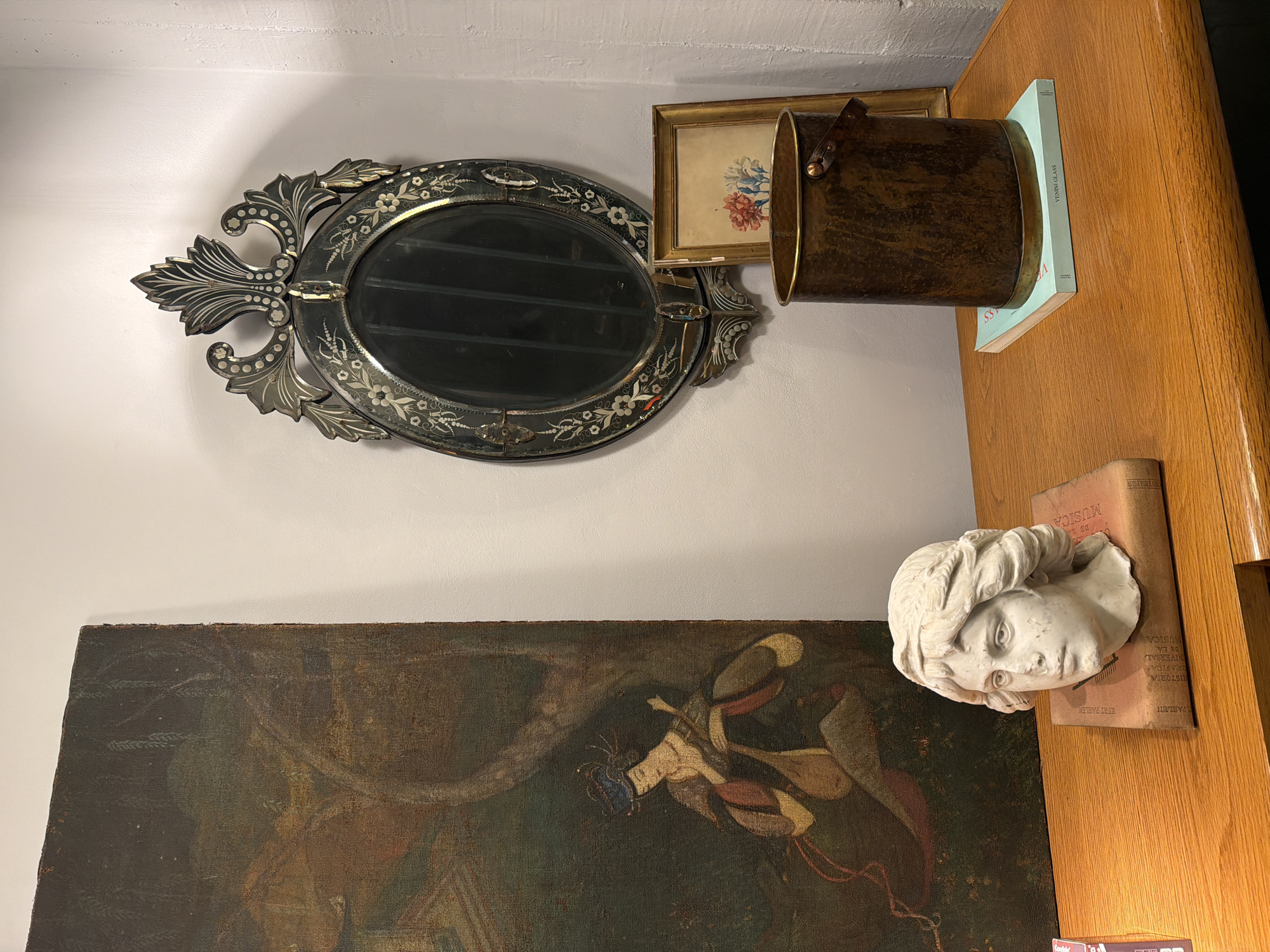 A still life arrangement is presented against a white wall, featuring a diverse collection of items. On the left side of the wall hangs an ornate oval mirror with a dark frame adorned with floral patterns. To the right of the mirror is a small, framed painting with floral motifs. A cylindrical metal container rests partially on the frame of the painting, propped up by a book with the title "VENINI GLASS" printed on its spine, resting atop a wooden surface.
Below the mirror and extending towards the left side of the image is a painting with a dark, textured surface, depicting figures in a muted color palette. On the wooden surface to the right, a white plaster bust of a head is placed atop a book. The book has text that reads "KURT PARLEN ISTORIA BASICA NIVERSAL DE LA USICA DE LA MUSICA".
The scene does not provide enough visual information to pinpoint a specific location in Buenos Aires, Argentina.