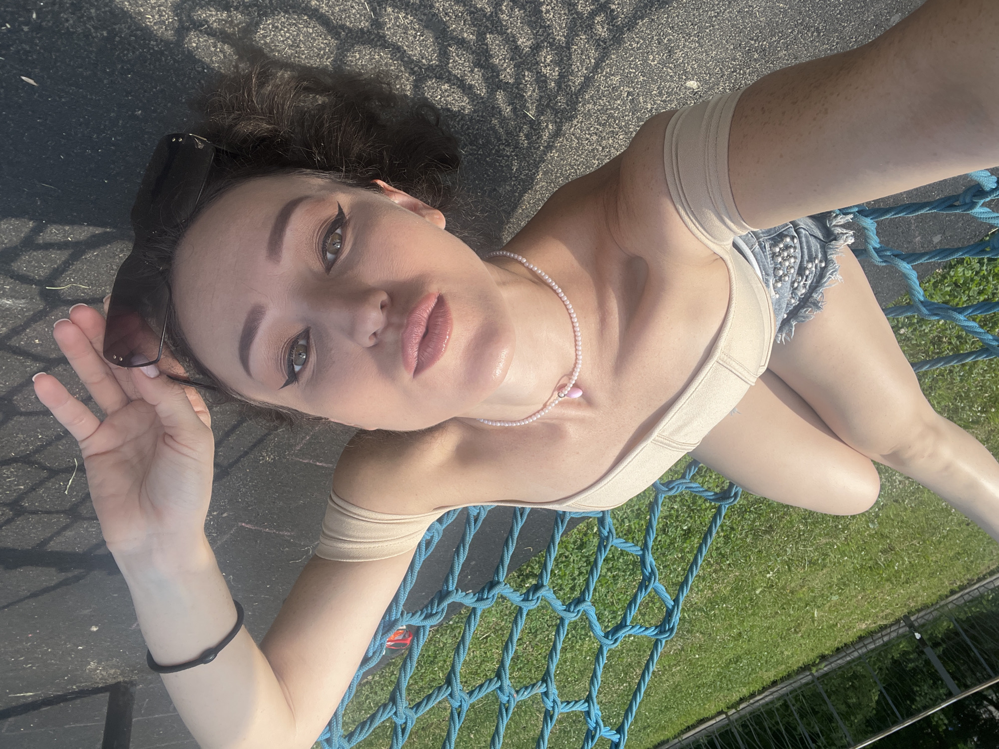 The image shows a young woman lying on a blue net. She is wearing a light-colored top and denim shorts adorned with silver studs. A pearl necklace with a pink pendant is around her neck. Sunglasses are perched on her head, and a black bracelet is on her left wrist. Her right hand is raised, with painted nails.
The scene appears to be outdoors, with a paved surface and grass visible in the background. The lighting suggests it is daytime, with shadows cast by an unseen object, possibly a tree or structure. The woman has a playful expression, with her head tilted and eyes looking towards the camera. The setting is likely a park or outdoor recreational area, given the net and grassy surroundings. There is no visible text in the image. The context indicates the location is Kazan, Russia.