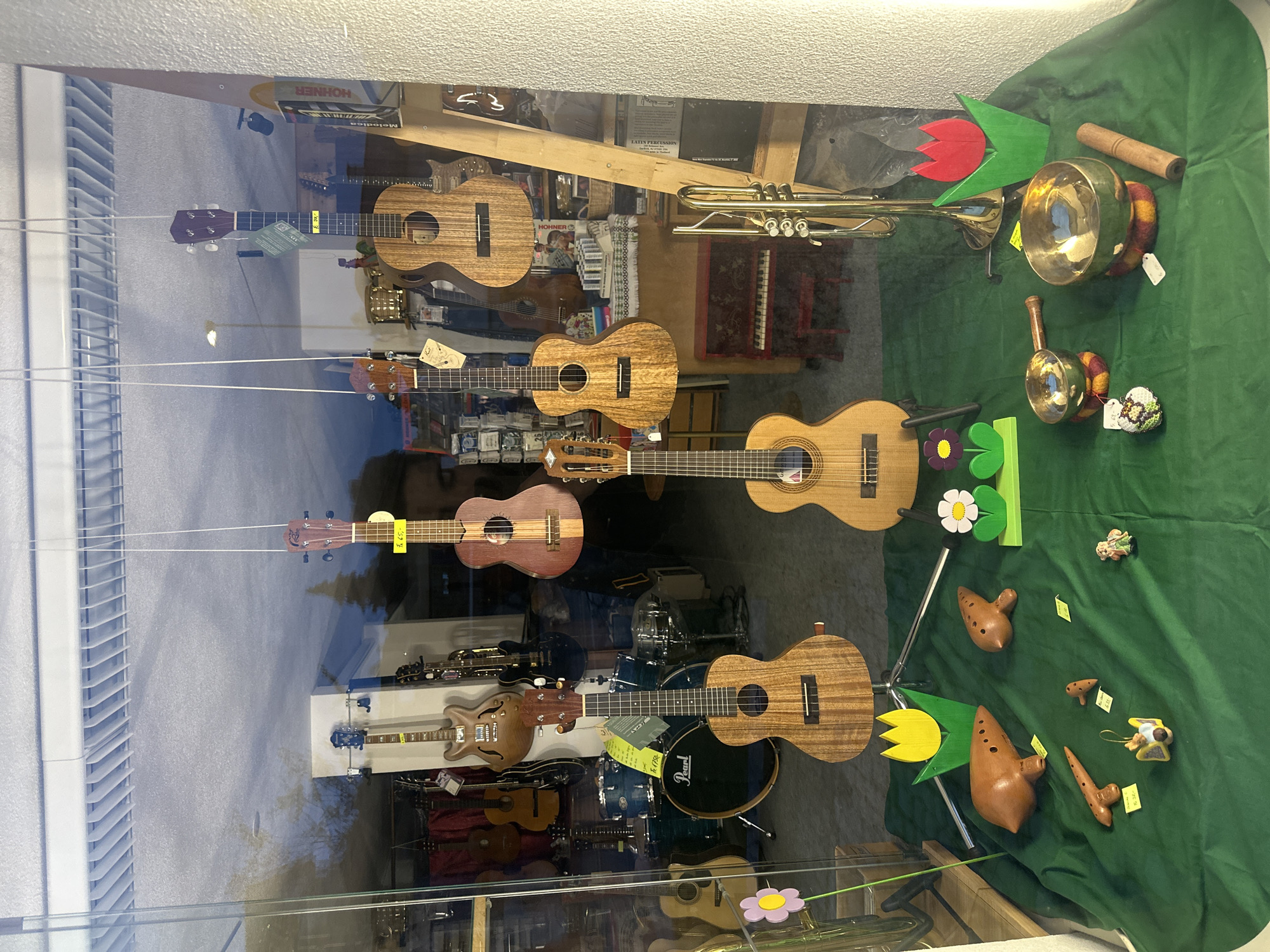 The image depicts a shop window display in Wald (ZH), Switzerland, showcasing a collection of musical instruments. Several ukuleles are suspended vertically from the top of the window by strings. Behind the ukuleles, a trumpet and other instruments such as bells and ocarinas are displayed on a green cloth-covered surface. Additional instruments, including electric guitars and a drum set, are visible in the background. The reflection on the shop window shows the street and surrounding buildings.