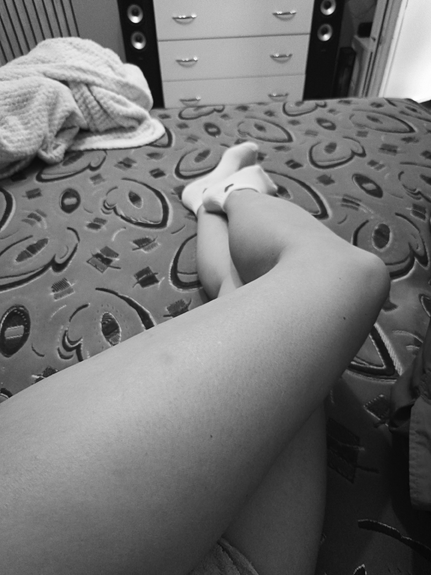 A black and white, medium shot photograph shows a person's crossed legs on a bed. The legs are covered in white socks. A patterned bedspread covers the mattress. A folded blanket sits on the left side of the bed. In the background, a light colored dresser with several drawers is flanked by two black speakers. The location is not evident in the photograph and it is not possible to determine if it is Sevastopol, Ukraine.