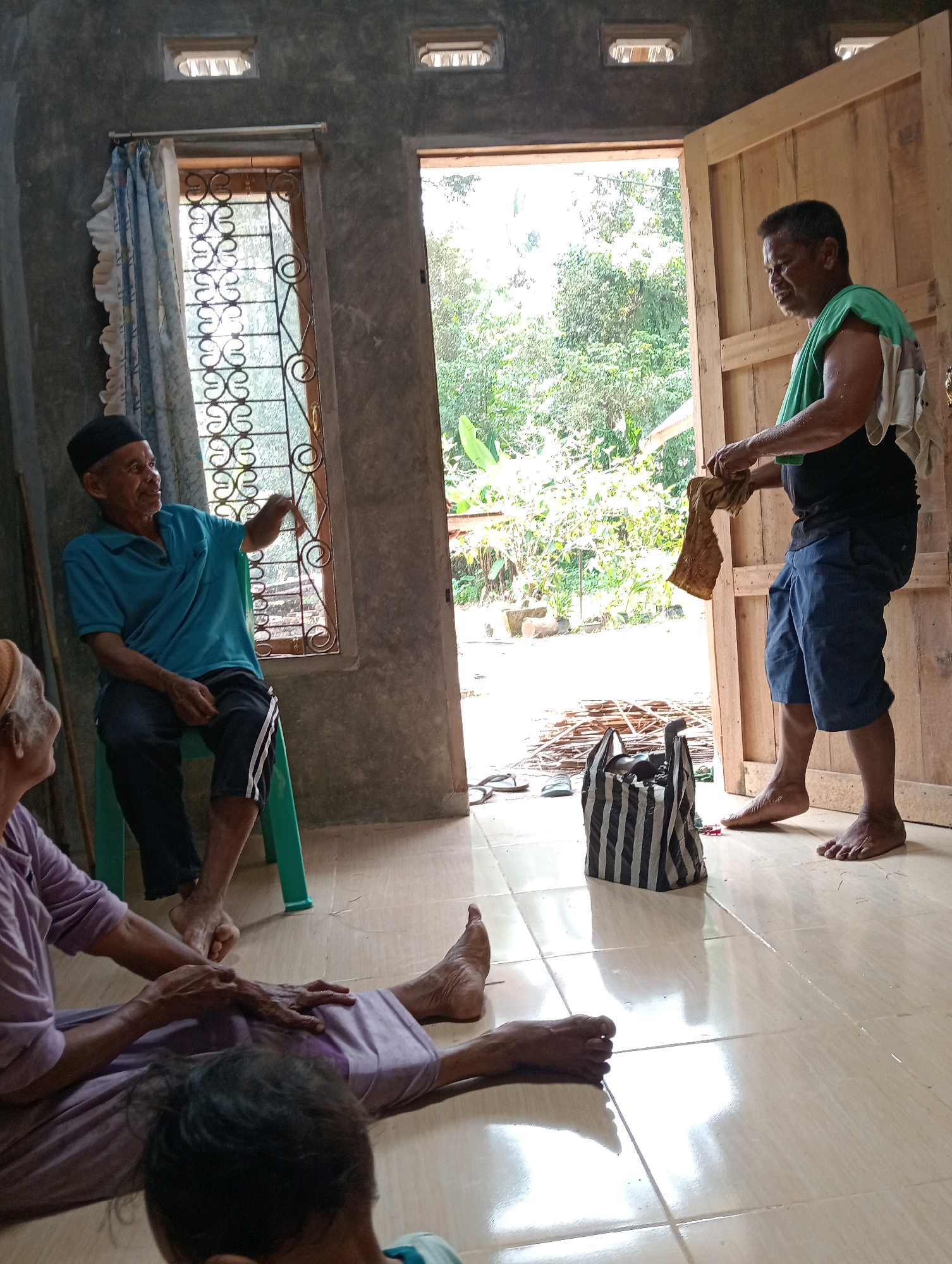 The image depicts an indoor scene with an open doorway leading to an outdoor area. In the foreground, two individuals are seated on the floor. One person is lying down, and another is seated, appearing to be attending to the person lying down. Their clothing is light purple.
In the middle ground, to the left, a man wearing a blue polo shirt and a black skullcap is seated on a green chair, looking towards a window. The window has a decorative metal grill and a light blue and white patterned curtain. To the right, a man in a black t-shirt and blue shorts, with a green towel draped over his shoulder, is standing in the doorway, holding a piece of fabric. A striped bag stands on the floor near him.
The environment appears to be a simple, rustic interior with concrete walls and a tiled floor. The outdoor view through the doorway shows lush green vegetation. The overall impression is one of a casual, possibly domestic gathering.
