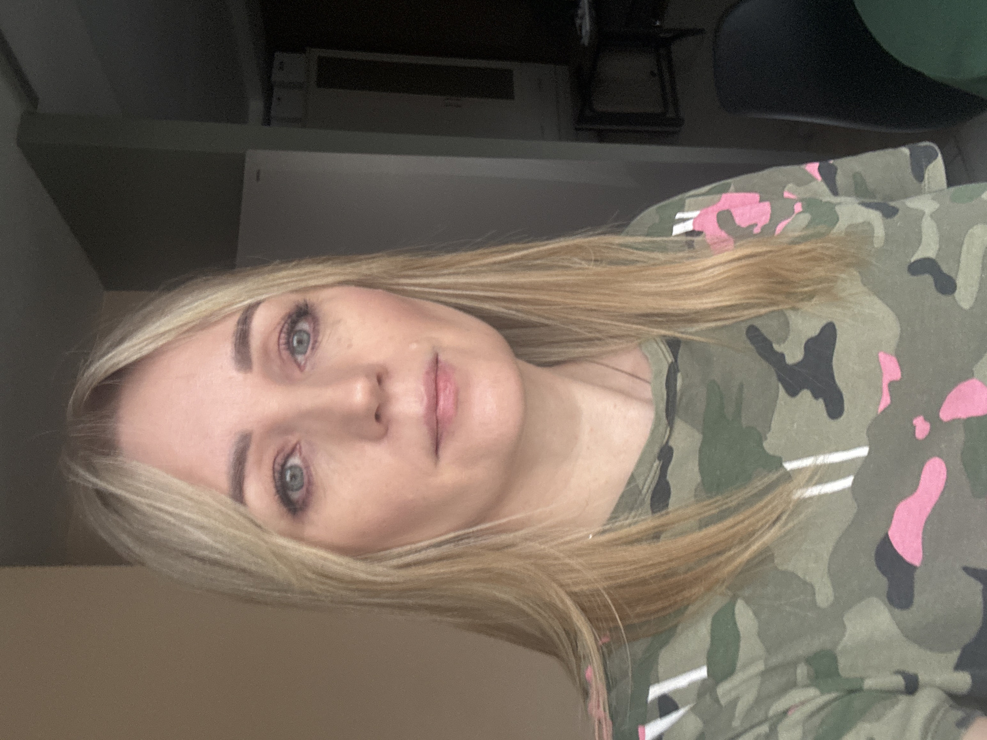 A woman with blonde hair and light-colored eyes is centered in the frame. She is wearing a camouflage-patterned shirt with pink and white accents. The background is a mix of blurred indoor elements including a shelf, a door, and furniture, suggesting a residential or office interior. There is nothing in the image that suggests the city of Votkinsk, Russia.