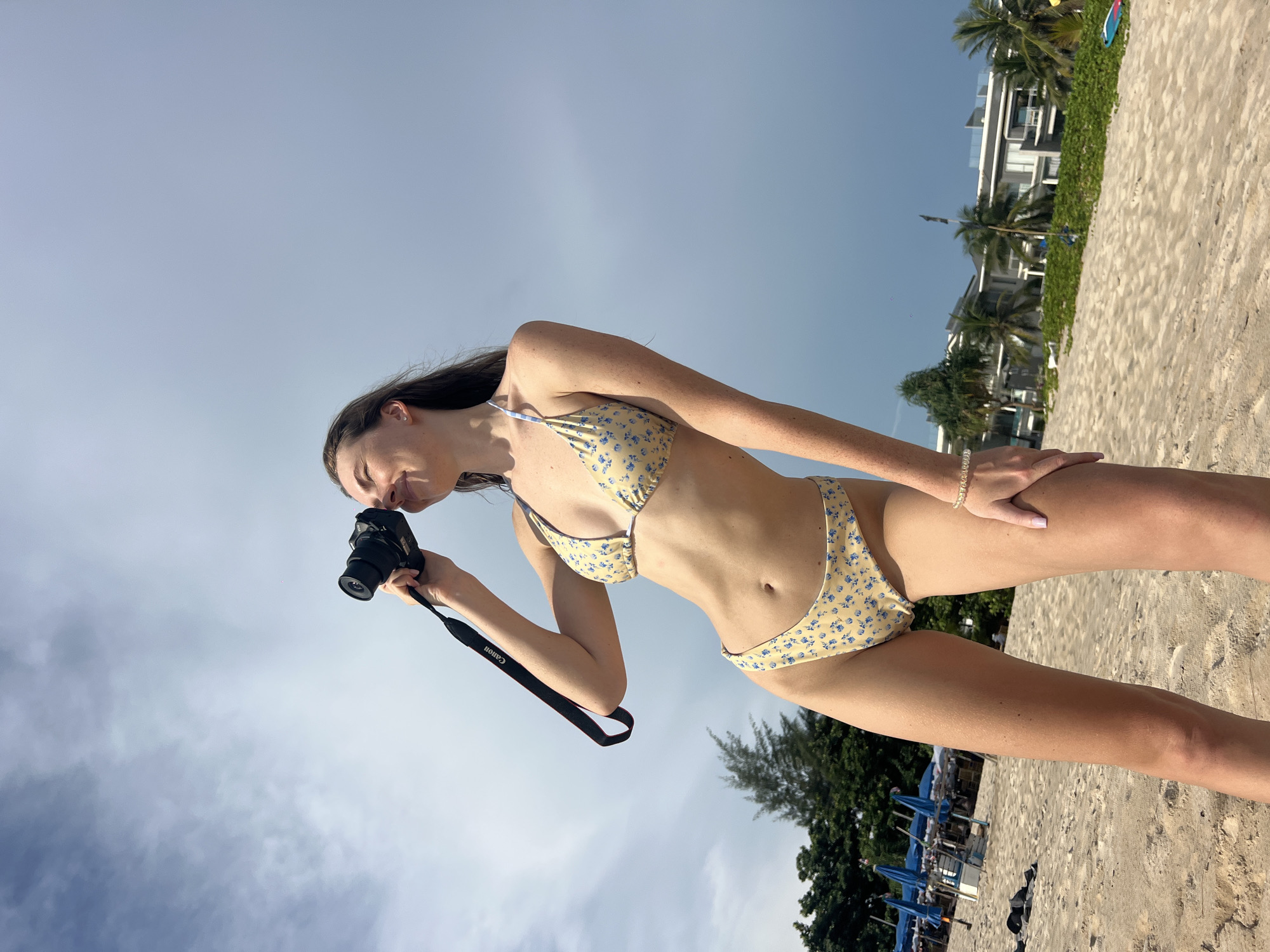 A woman wearing a yellow floral bikini stands on a beach, holding a black Canon camera up to her eye. The beach is sandy, with some vegetation and trees visible in the background. Buildings and palm trees are also visible. The sky is blue with some clouds. The location is likely in Choeng Thale, Thailand.