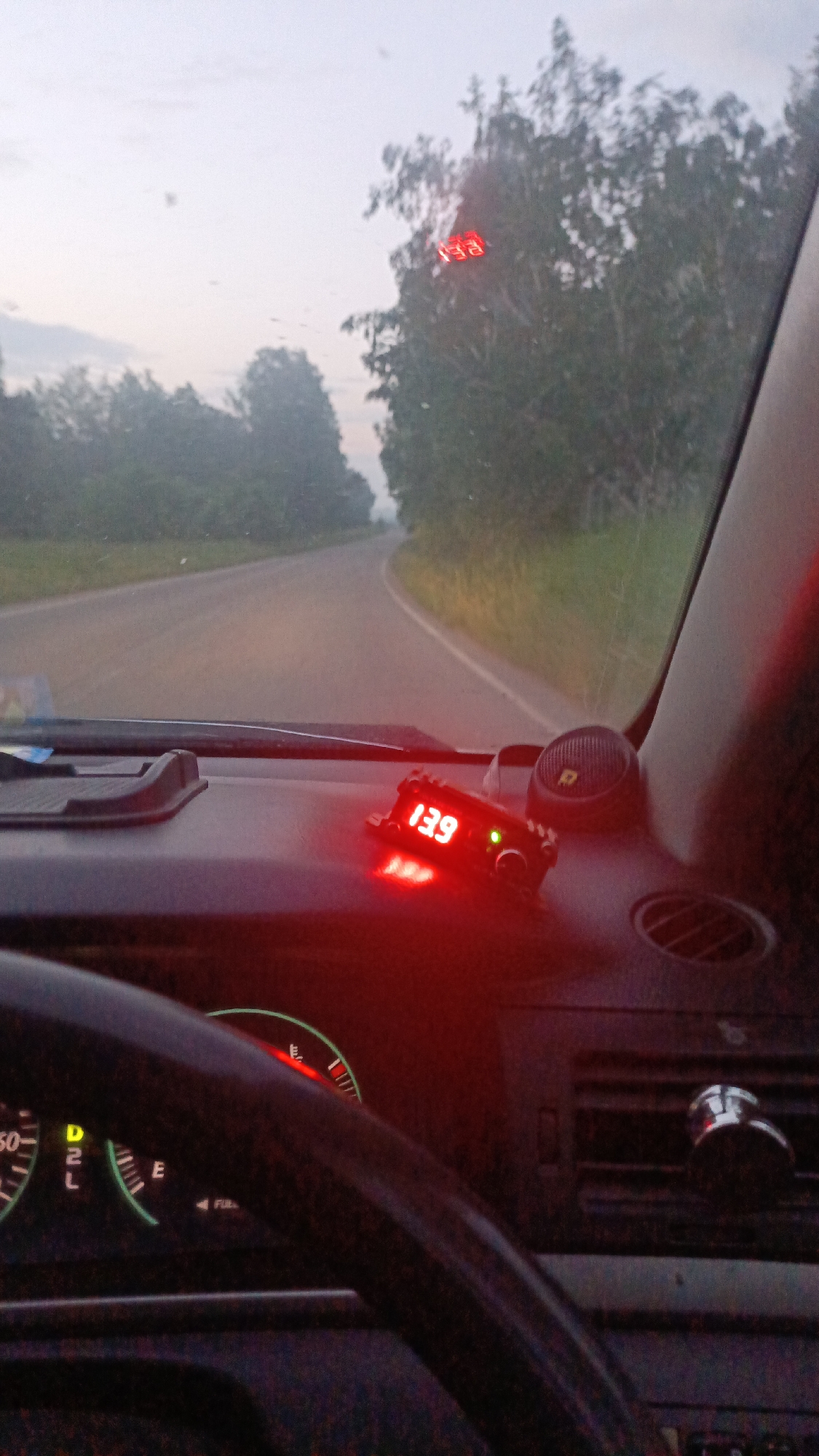 The image shows the interior of a car driving on a road with trees on either side. The dashboard has a red digital display showing "13.9". There are also instrument cluster gauges visible, including a tachometer with a red needle pointing upwards and a fuel gauge. The sky is overcast. The location is identified as Zuy, Russia.