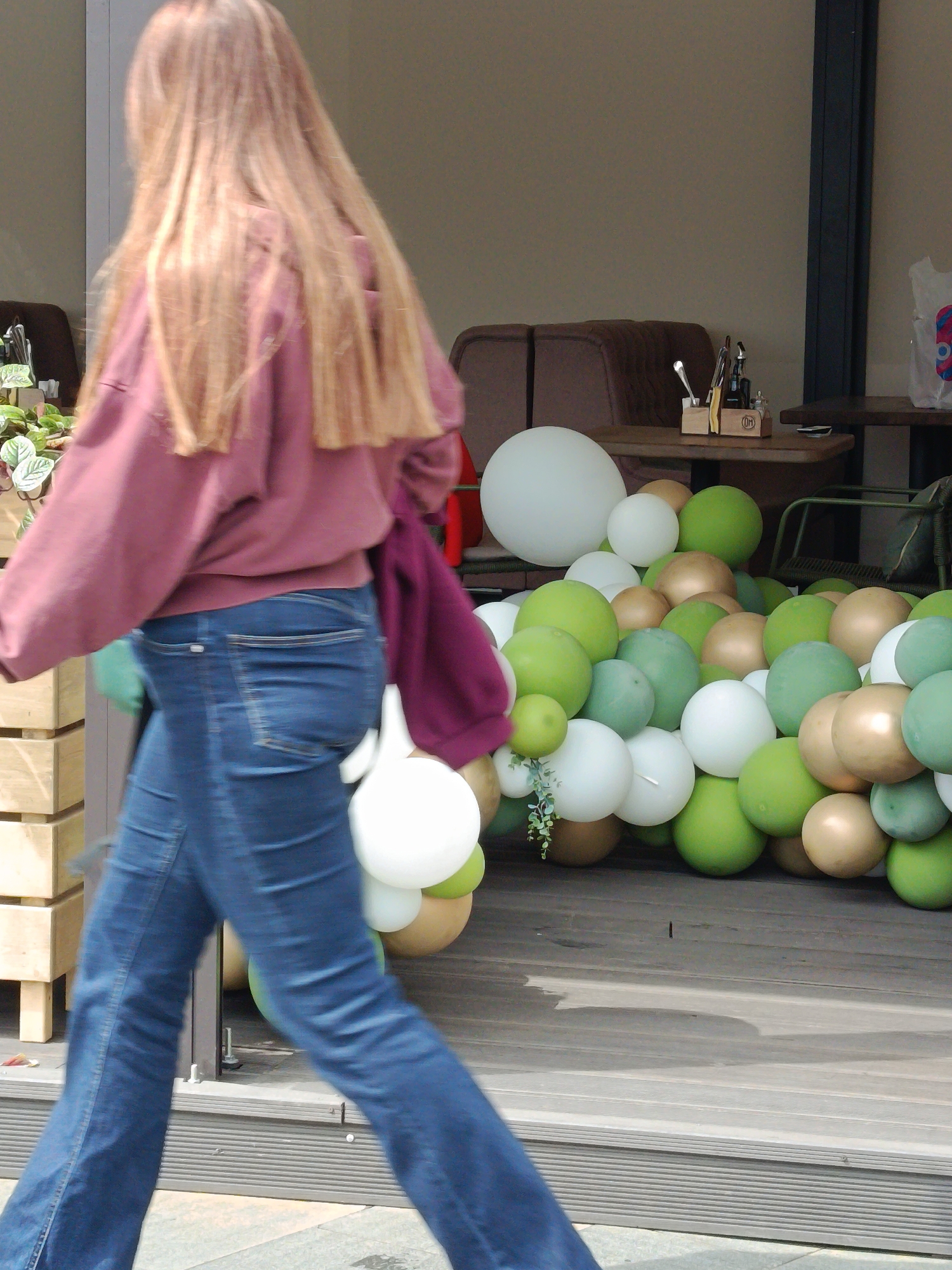 A photograph captures a woman in motion, walking away from the camera. She has long, light brown hair and is wearing a mauve sweatshirt and blue jeans. Her left arm obscures the view somewhat, but she seems to be carrying white balloons and a magenta tote bag. She is walking past an outdoor cafe-like space, decorated with a large arrangement of green, white, and gold balloons. A wooden planter box and a dark wooden table with a caddy holding cutlery are also visible. Based on the style of the architecture, location, and decorative elements, this scene might be located in the city of Moscow, Russia.
