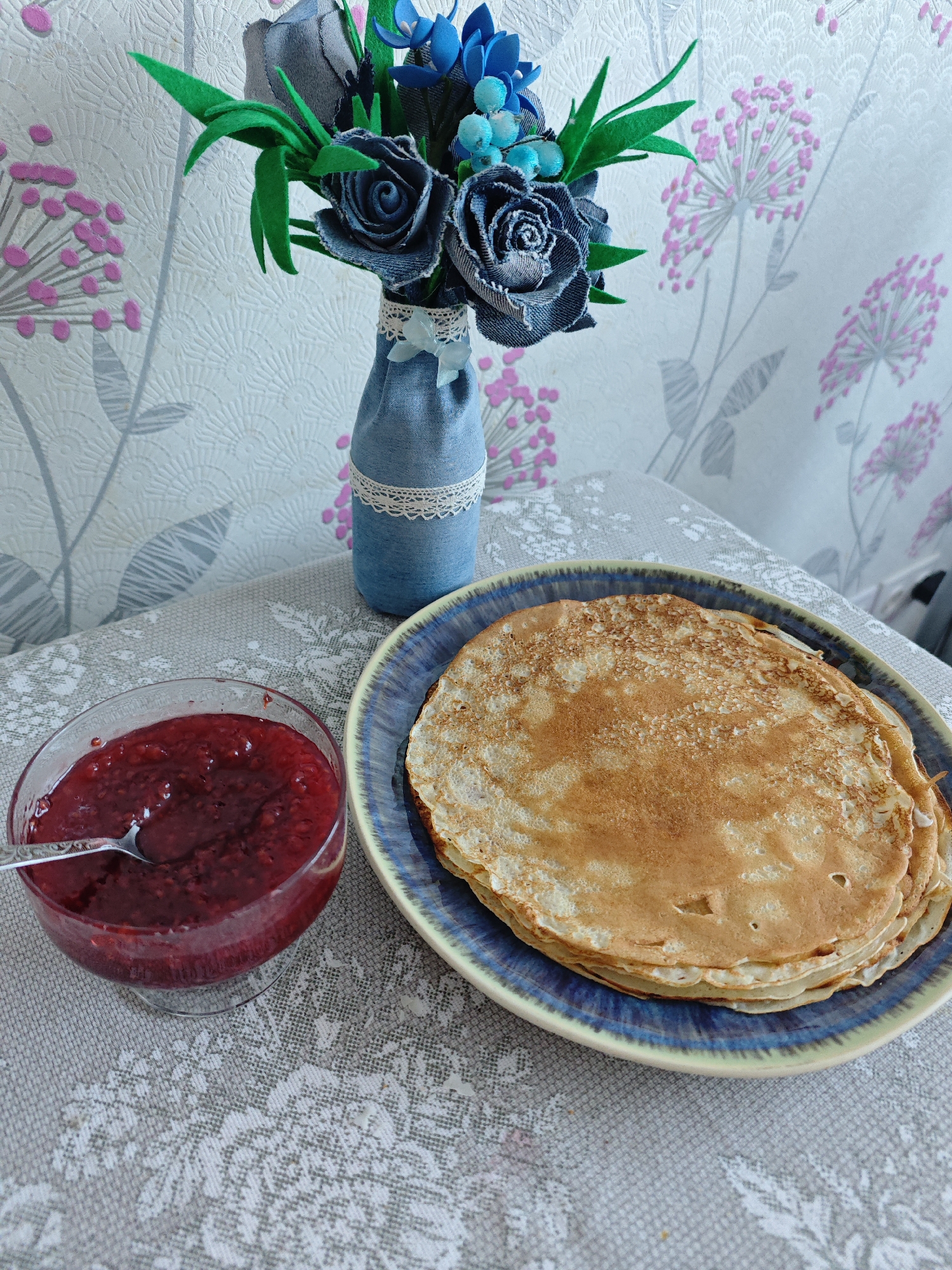 The image depicts a still life arrangement of food and decorative items, likely indoors. The scene includes a plate of stacked pancakes, a glass bowl filled with red fruit preserve (possibly raspberry or similar), and a small denim-covered vase holding a bouquet of fabricated flowers. A spoon rests within the preserve. The arrangement is situated on a gray tablecloth with a floral design. The background features a wall covered in wallpaper with a floral pattern in pink and gray. The location may be in the city of Markovskii, Russia. There are no people visible in the image.