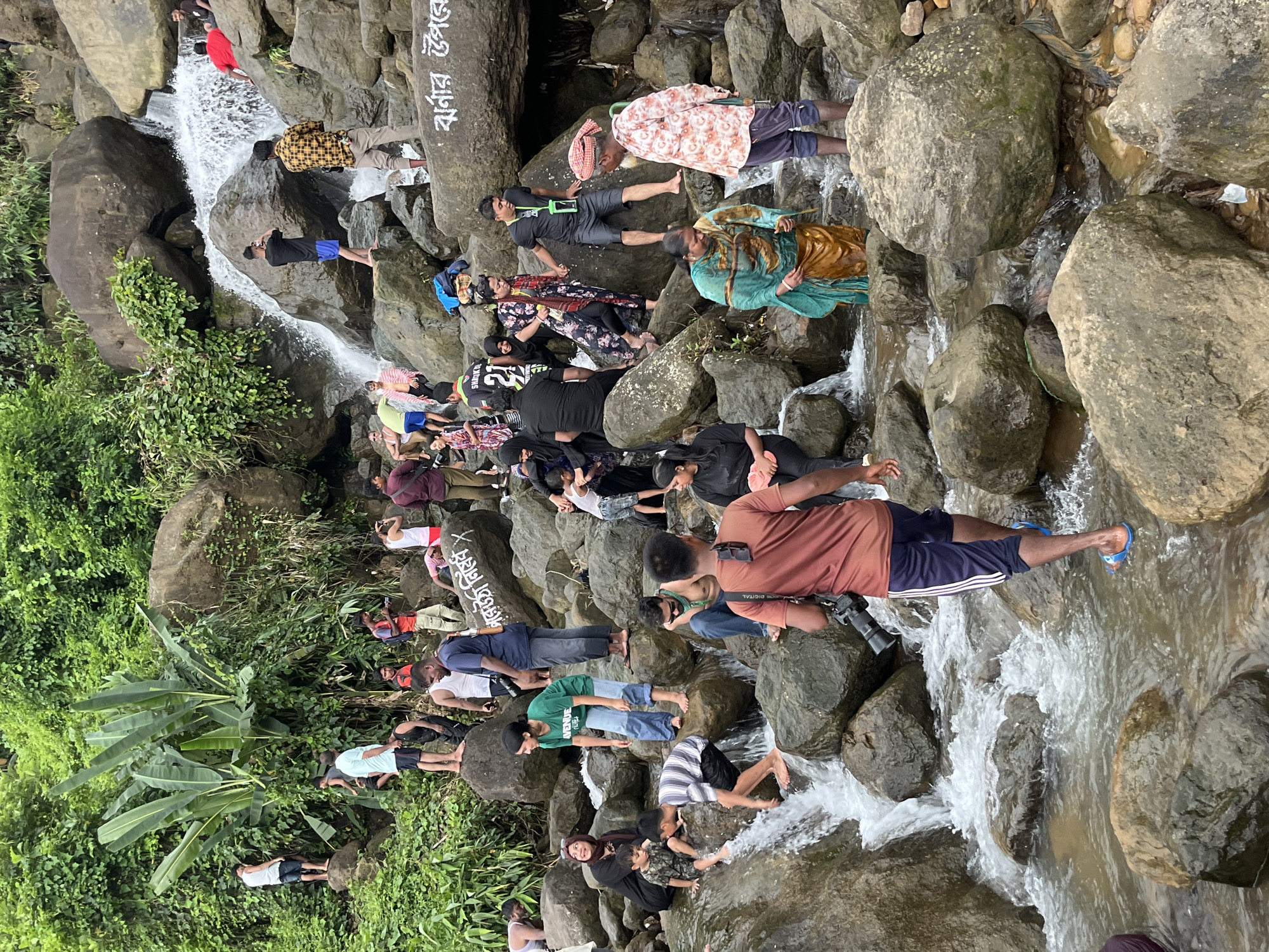 The image depicts a lively scene at a waterfall in Jaflong, Bangladesh, where numerous people are enjoying the natural surroundings. A group of individuals, dressed in a variety of casual attire, are positioned around the cascading water and on the surrounding rocks. Several are wading in the shallow water, while others are perched on the stones, some taking photographs with their phones or cameras. The lush green foliage and large boulders characteristic of Jaflong's landscape dominate the background. The atmosphere appears to be one of recreation and community engagement, with people of different ages interacting and engaging in activities such as swimming and photography.