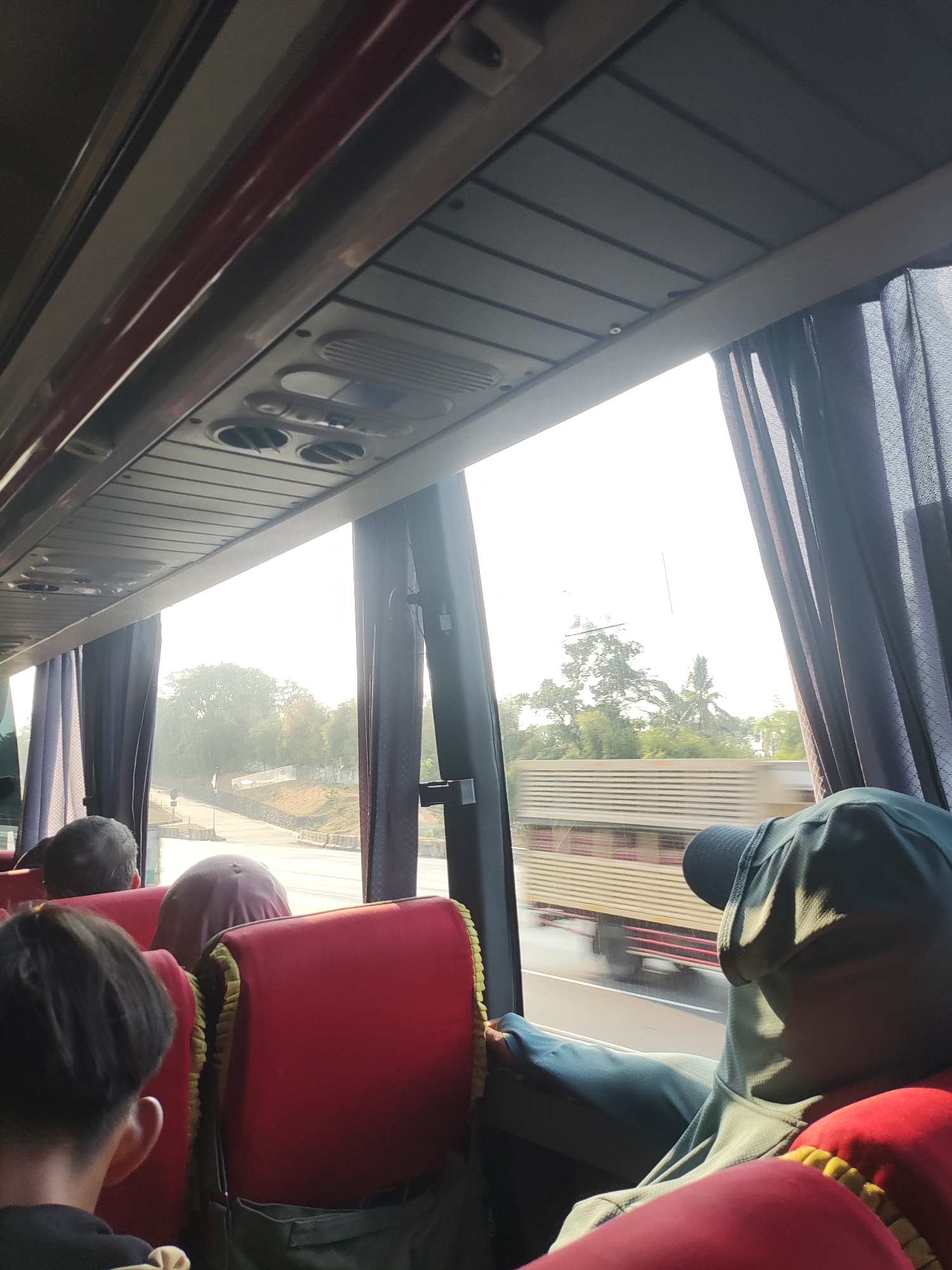 Inside a passenger bus, several people are seated. The focus is on the interior of the bus, with a view of the exterior through the windows. The bus is in motion, with a road and trees visible outside. A large truck is passing by on the road. The seats are red with yellow trim. The ceiling of the bus has air vents and lights. The image appears to have been taken during the day, with bright sunlight streaming in from the windows. The general atmosphere is one of travel and transit. The location is identified as Karawang, Indonesia, implying this is a bus traveling through that region.