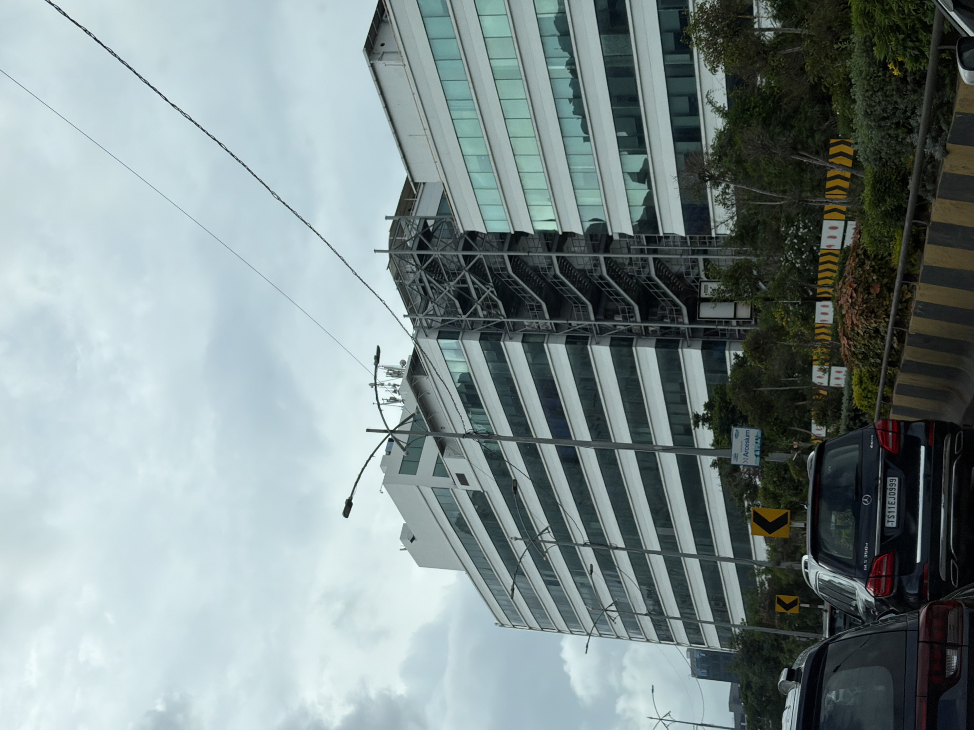 The image shows a daytime scene in Hyderabad, India, with modern office buildings and traffic. In the foreground, a dark-colored SUV is visible, its license plate reading "TS11EJ0999". Behind it, a Mercedes-Benz GL S 350d is also parked. The buildings are multi-story structures with white facades and rows of tinted glass windows. One building features an external metal staircase. The sky is overcast with grey clouds. Road signs indicating a sharp left turn are present. A sign with "Arcelsium" is also visible. Greenery, including trees and bushes, lines the roadside. The overall impression is of an urban environment with ongoing vehicular traffic.