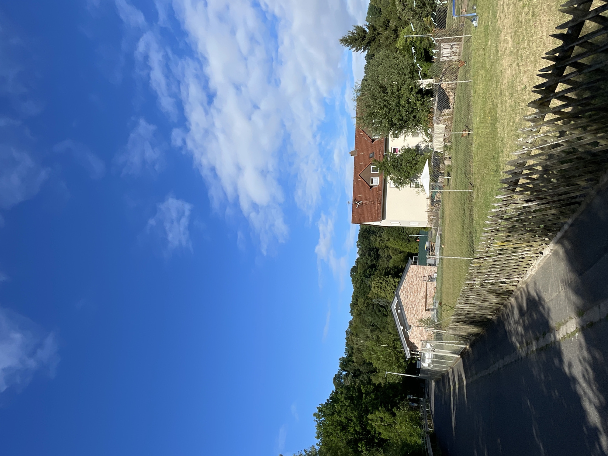 The image depicts a rural scene in Neukirchen, Germany, on a clear, sunny day with scattered white clouds in a bright blue sky. In the foreground, a paved road runs diagonally from the bottom left towards the center of the frame, casting shadows from unseen objects. Adjacent to the road, a rustic wooden picket fence runs along the right side of the image, separating the road from a grassy area. Beyond the fence, a small, brick building with a grey roof is visible, and further back, a larger, light-colored building with a red tiled roof stands out. The area behind the fence appears to be a garden or yard with some trees, a white umbrella-like structure, and a few chickens grazing on the grass. A dense line of trees forms a backdrop across the middle ground and extends into the background on the left and right sides of the frame. The overall impression is one of a tranquil, semi-rural environment.