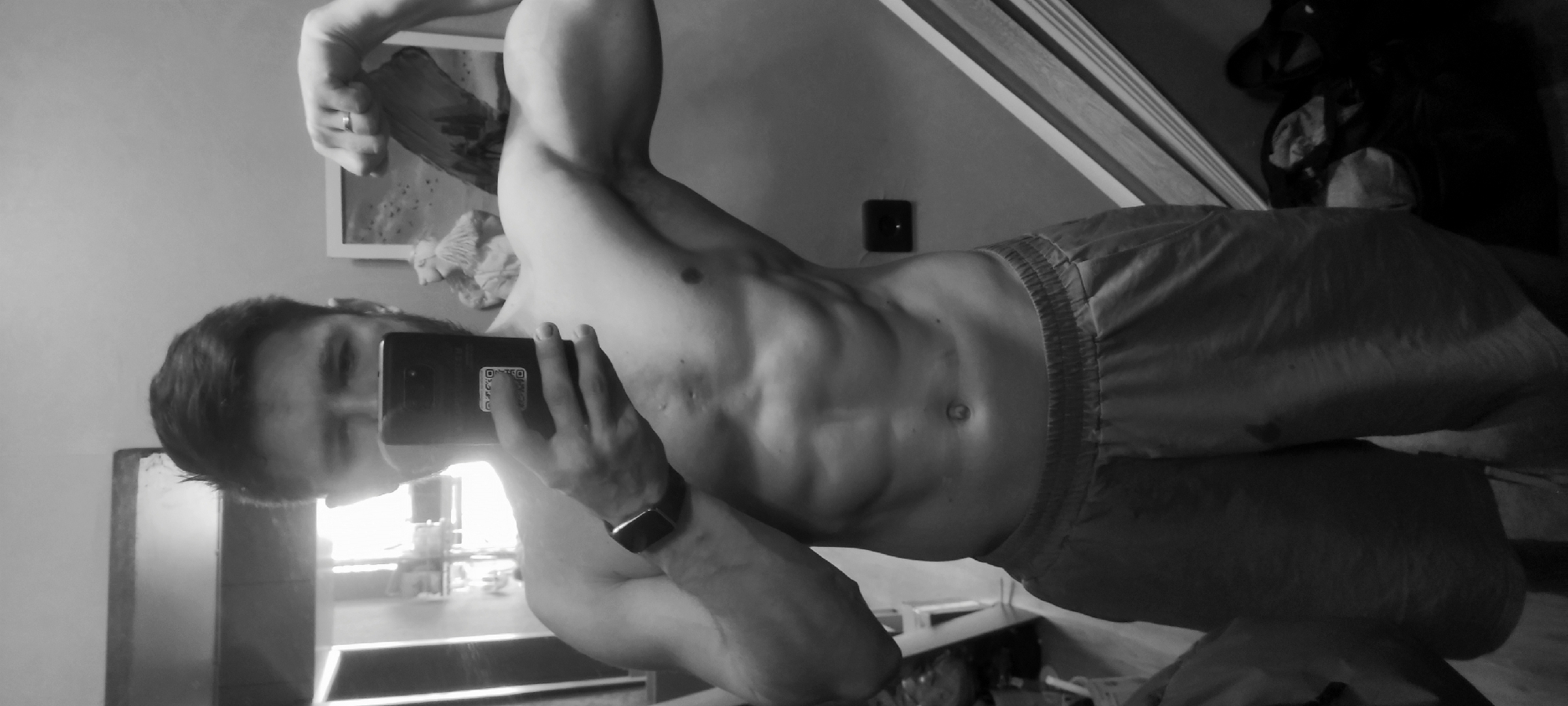 A shirtless man wearing shorts is taking a selfie in a mirror. The man is muscular with defined abdominal muscles and biceps. He is holding a smartphone in his left hand, covering part of his face. The smartphone has a QR code visible on the back. He is wearing a smartwatch on his left wrist. The room appears to be a bedroom, with a window on the left side of the frame and a shelf or cabinet visible behind the man. There is a picture frame on the wall. The image is in black and white. The location is not discernible.