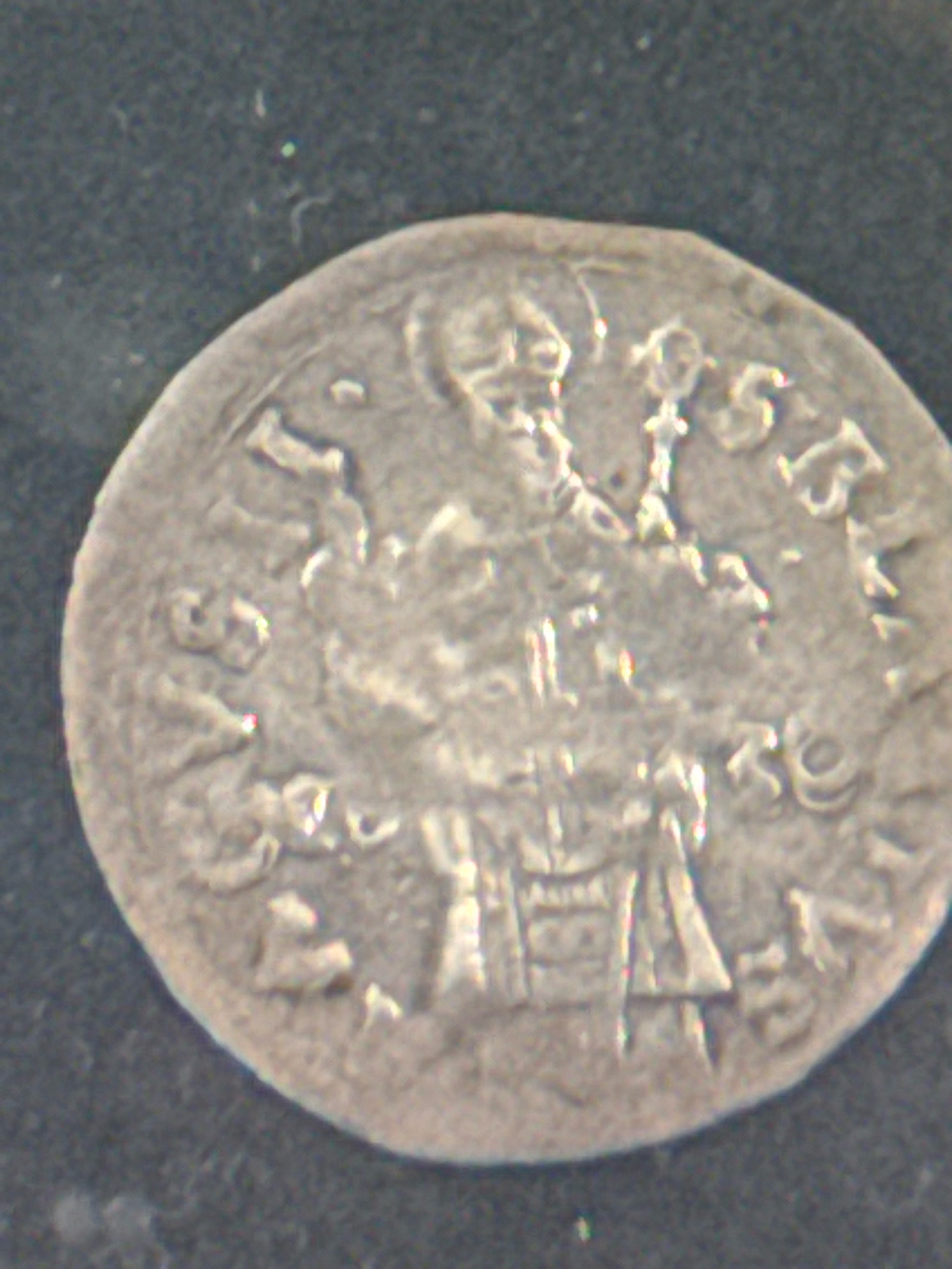 The image is a close-up, high-angle shot of a silver coin, likely an ancient or medieval artifact, lying on a dark, textured surface. The coin is round with a slightly irregular edge and exhibits significant wear, obscuring many of the finer details.
The obverse of the coin appears to feature a central motif, possibly a stylized building or a coat of arms, surrounded by inscriptions. The inscription on the top right portion of the coin seems to contain numerals that might represent a date, with visible elements resembling "5" and "5". The inscription on the left side of the coin is partially obscured and worn, but some characters can be discerned. The overall impression is of a historically significant object with a weathered surface.
This coin is not located in the city of Dubrovnik, Croatia. It is a standalone object depicted in a close-up photograph.