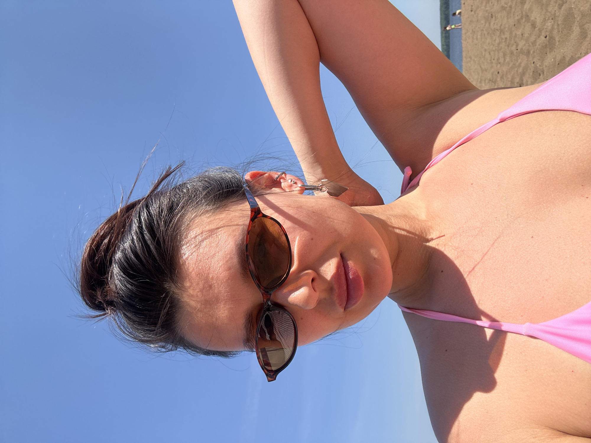 A woman wearing a pink bikini and sunglasses is photographed from a low angle, with a blue sky in the background. Her hair is pulled back. The location appears to be a beach in Chaikovsky, Russia. In the background, the beach and the sea can be seen.