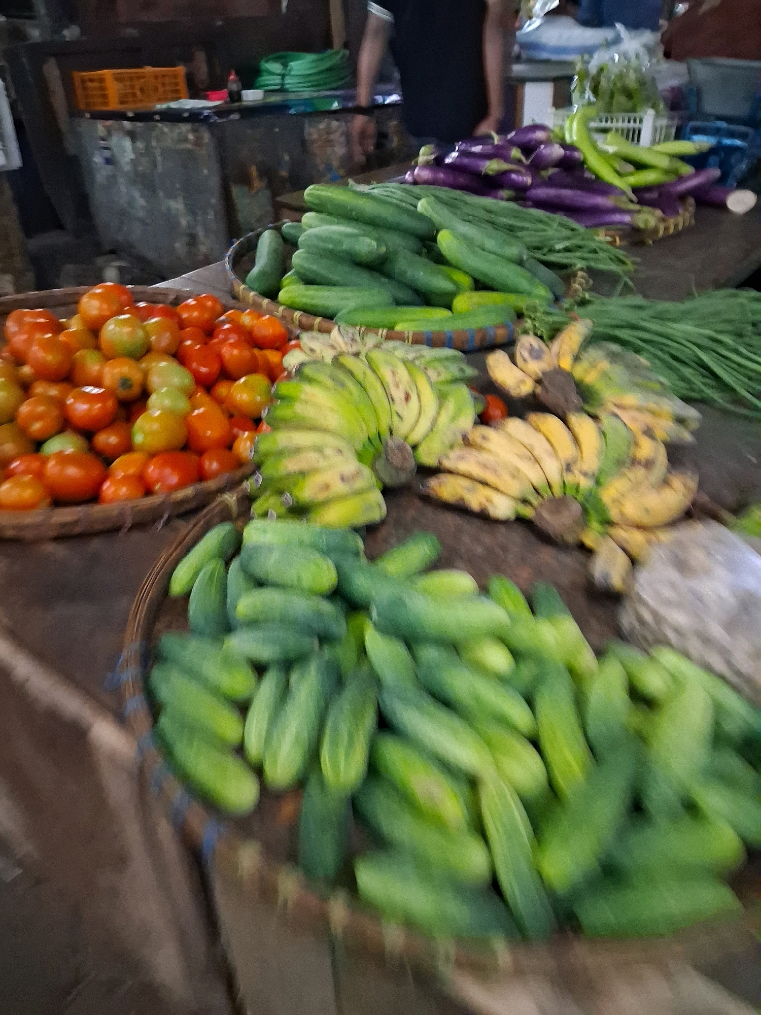 A low-angle, close-up shot captures a market stall in Surakarta, Indonesia, showcasing a vibrant display of fresh produce. The foreground is dominated by a woven basket overflowing with numerous bright green cucumbers. To the left, another basket holds a large quantity of red and orange tomatoes. Behind these, a bunch of ripe yellow bananas with brown spots is visible, alongside green onions and what appear to be green peppers or chilies. In the background, dark purple eggplants are piled high. The stall is set against a rustic backdrop of a weathered counter and some indistinct shelving units, with a person's dark clothing visible in the upper right corner. The lighting is somewhat dim, suggesting an indoor or shaded outdoor market setting.