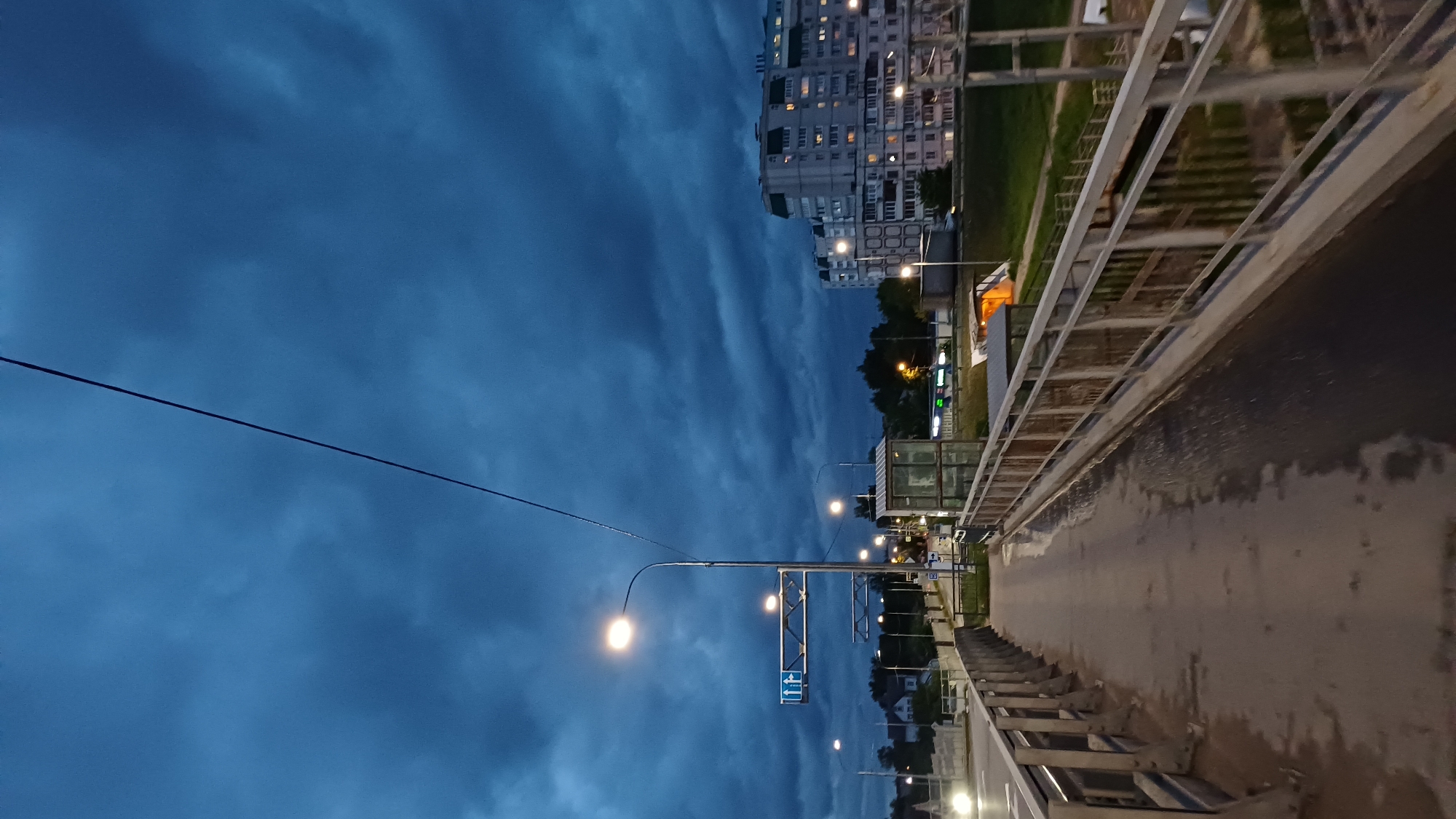 The image depicts a nighttime scene on a bridge in Obukhovo, Russia. The view is oriented sideways, with the city skyline and a dramatic, cloudy sky on the left side of the frame. A large apartment building with illuminated windows is visible in the distance. Streetlights line the bridge, casting a warm glow on the wet pavement and metal railings. A power line stretches across the sky. A bus stop sign is also visible. There are no people visible in the image. The focus is on the urban landscape at dusk.