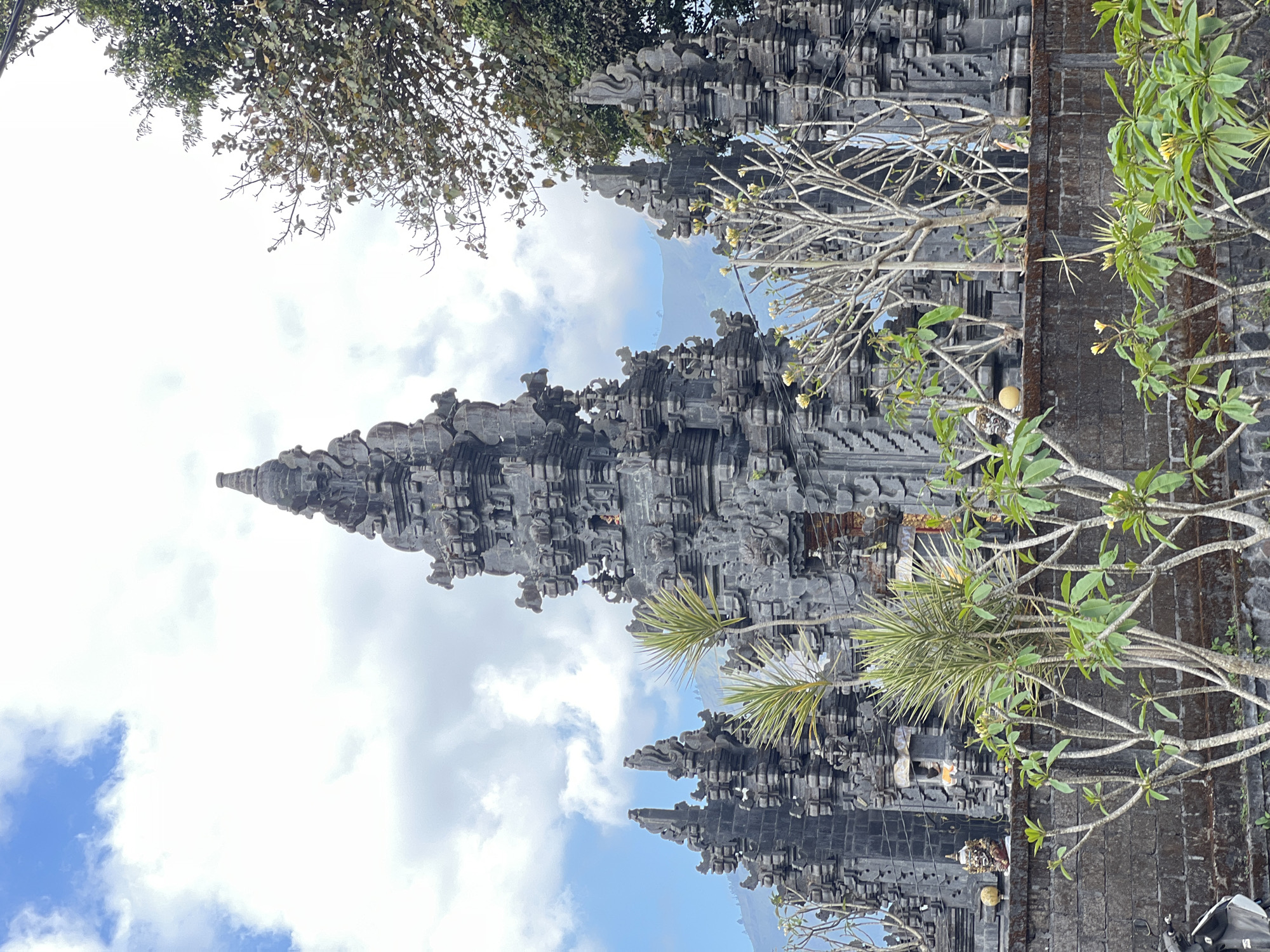 A stone temple structure with intricate carvings is shown in the city of Kintamani, Indonesia, against a backdrop of a bright sky with scattered clouds. The temple has a tall, tiered central spire and several smaller flanking structures. In the foreground, lush green plants with some flowering branches are visible, with a stone wall bordering the temple grounds. A portion of a motorcycle can be seen on the left side of the frame. The overall impression is one of architectural grandeur set within a tropical landscape.