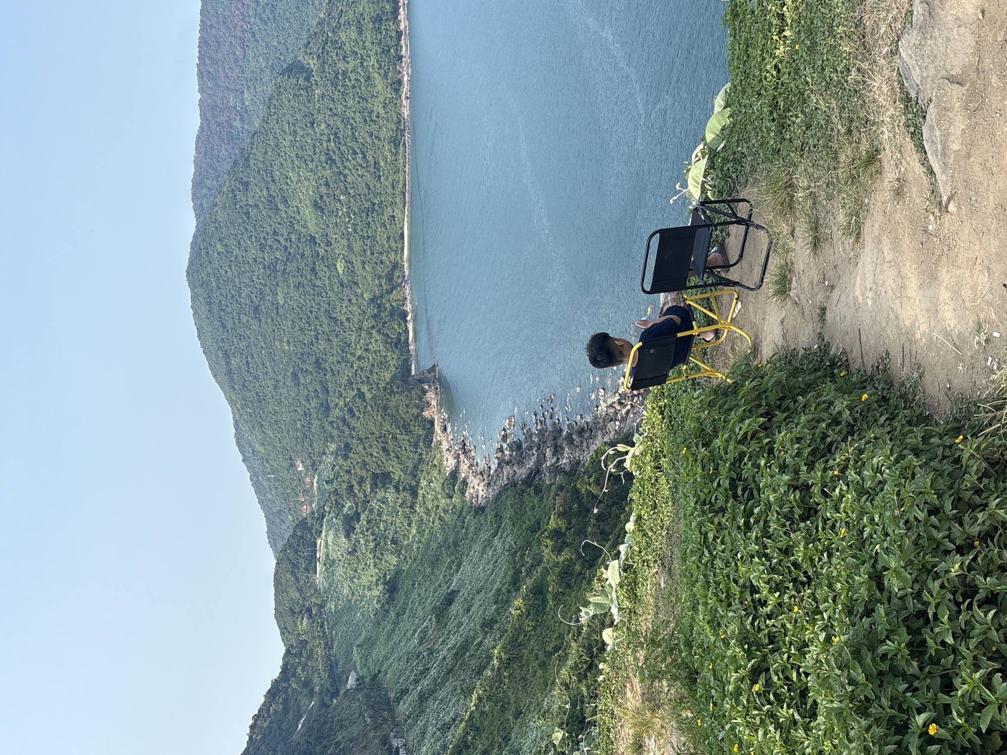 A person sits in a yellow-framed chair on a grassy hill overlooking a body of water and a green, mountainous coastline. The person is facing away from the viewer and appears to be looking at something in their lap. Another black metal chair is placed behind the person, further up the hill. The hill is covered in vegetation, including some flowering plants. The body of water is blue and stretches to the horizon. The mountainous coastline is green and lush. The sky is light blue and clear. The location is likely Son Tra Peninsula in Đà Nẵng, Vietnam, given the topography and coastal features.