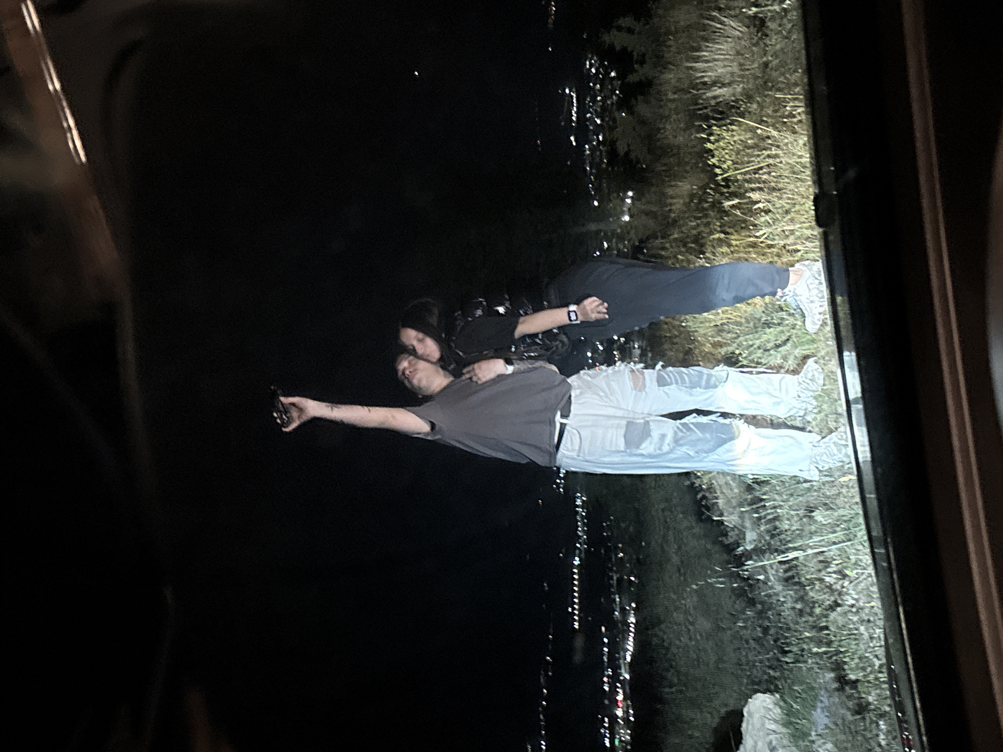 A nighttime image taken from inside a vehicle, likely a car, looking out a window at a couple. The couple, a man and a woman, are standing near a body of water or a dark expanse, possibly a river or lake, with lights reflecting in it, creating a cityscape backdrop. The man, wearing a gray shirt and light-colored, ripped jeans, is holding a device in his outstretched left hand, possibly taking a photograph or video. The woman is wearing darker clothing and is leaning in towards the man, with her head near his. The environment appears to be near or within Marulevo, Bulgaria, based on the reference information. The foreground, the interior of the vehicle, is dark. The image suggests the couple is capturing a moment near an urban area.