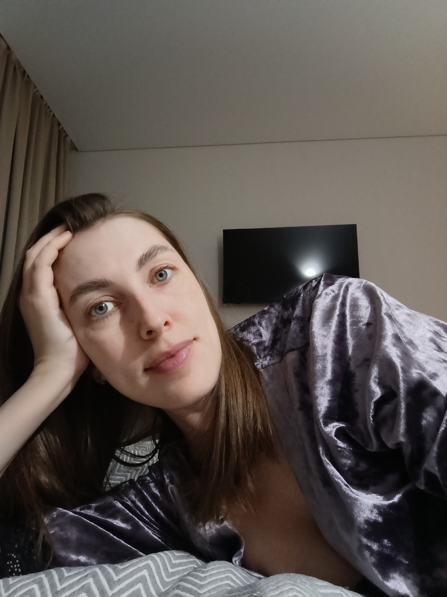 A close-up portrait features a young woman, likely in her 20s or early 30s, lying down and looking directly at the camera. She has fair skin, prominent blue eyes, and long, straight brown hair. Her left hand gently rests against her face, supporting her head, while a soft, subtle smile plays on her lips, conveying a relaxed and serene expression.
She is dressed in a plush, velvety garment of a muted purple or grayish-lavender color, which appears to be a robe or loungewear. The fabric exhibits a soft sheen and texture. She is positioned on a bed, indicated by a grey and white chevron-patterned duvet or blanket partially visible in the foreground.
The background reveals an indoor setting, characteristic of a bedroom or hotel room. Light brown or beige curtains are seen on the left side of the frame, and a black flat-screen television is mounted on a light-colored wall on the right. The lighting is soft and subdued, suggesting it is evening or nighttime. No text is visible in the image. This scene is set in Chaikovsky, Russia.