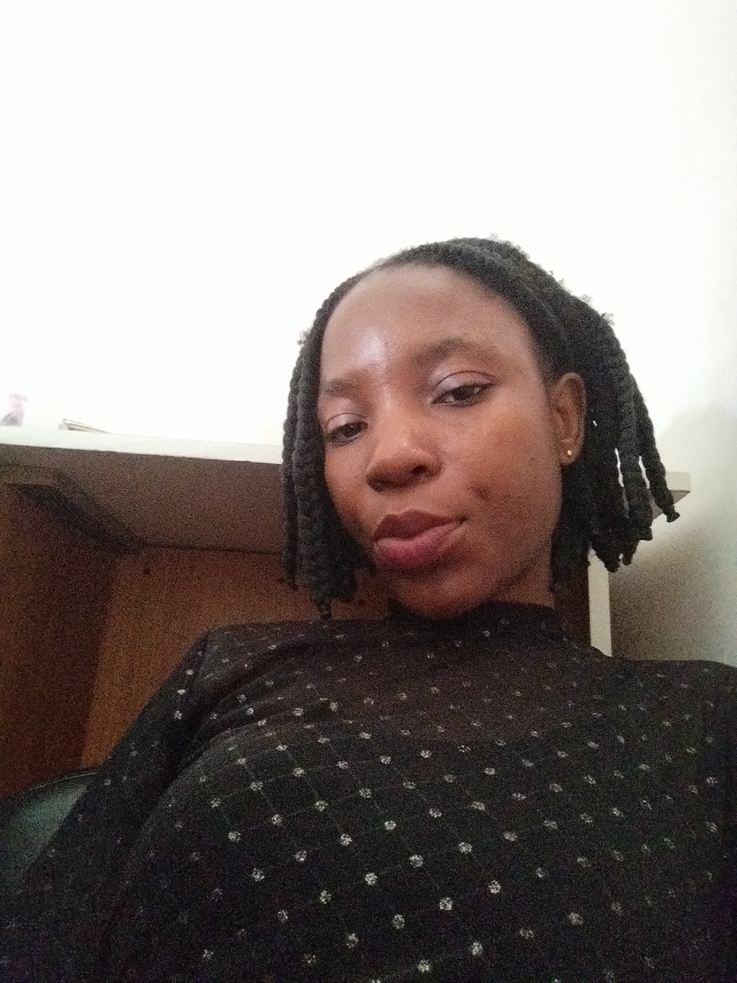 A medium shot taken from a slightly high angle shows a young Black woman with braided hair and a black, patterned top. She is positioned in the foreground against a plain white wall. To her left, a dark brown shelf unit is visible. The woman is looking towards the camera with her head tilted slightly. A small gold stud earring is visible on her right ear. The lighting appears to be artificial and is casting a slight shadow on the wall behind her. There are no other individuals or distinct objects in the immediate vicinity. The scene is indoors, with the background suggesting a domestic or office environment, possibly a room in Uselu, Nigeria.