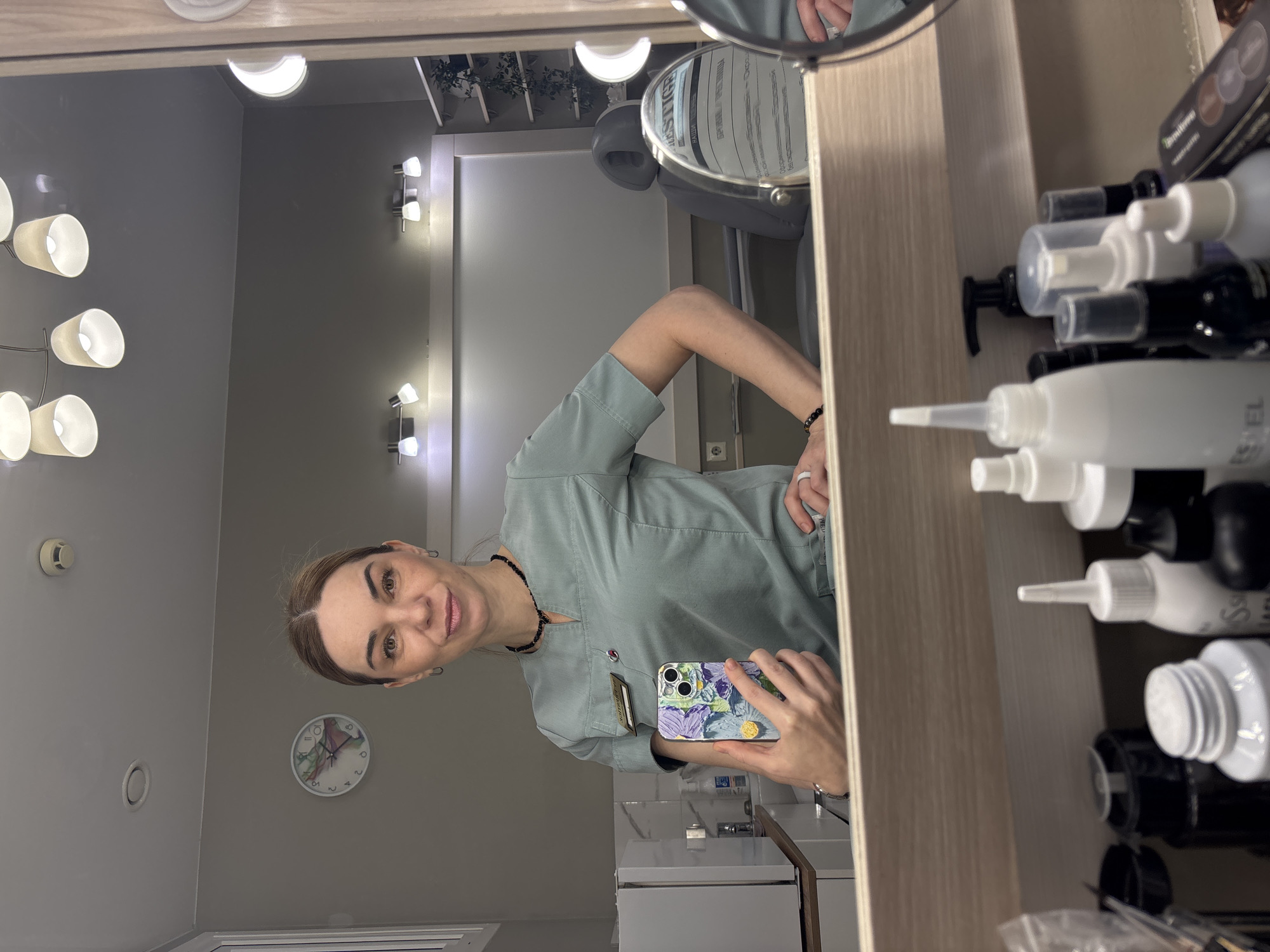 A woman is taking a mirror selfie in what appears to be a professional setting, possibly a salon, spa, or medical office. She has light brown hair, pulled back, and is smiling subtly at her smartphone held in her left hand. She wears a light green/teal scrub-like uniform, a dark beaded necklace, a white ring on her right ring finger, and a dark bracelet. Her phone case features a colorful floral pattern with a yellow PopSocket. A name tag is pinned to her uniform, but its text is unreadable.
The room has neutral grey walls and is illuminated by multiple modern wall sconces with white shades and a half-moon shaped ceiling light. On the right side of the mirror's reflection, a wooden shelf or cabinet holds numerous white, black, and clear plastic bottles of various products, some with pumps or nozzles. "ESTEL" is visible on one white bottle. Product boxes are also stacked on the shelf.
In the background, a white door is visible, along with a part of a professional chair, possibly a salon or treatment chair. A small, round magnifying mirror on a stand is prominent, displaying the word "PRESTIGE" and some Cyrillic text, including "НАШИ" (NASHI). A white wall clock with colorful abstract markings is on the left wall, and a smoke detector is on the ceiling. A green potted plant is reflected on a shelf in the upper right. The scene suggests a clean, well-lit, and organized professional environment.