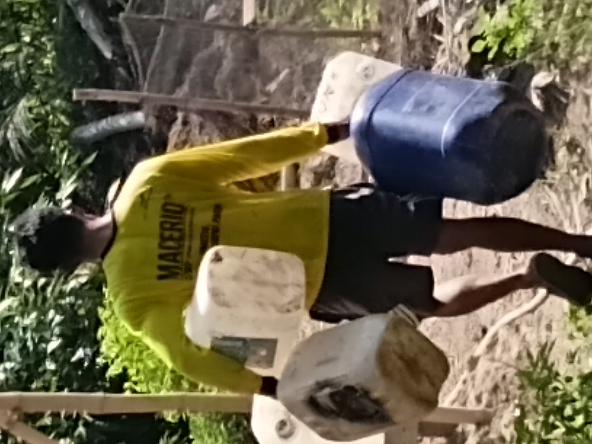 In Margo Rukun, Indonesia, a person wearing a yellow long-sleeved shirt with the text "MACERIO" visible on the back walks away from the camera. The person is carrying three large plastic containers: one white, cube-shaped container in their right hand, a larger, irregular-shaped white container in their left hand that is partially obscured, and a blue, barrel-shaped container in front of them, being held by their legs. The person is wearing dark shorts and dark flip-flops. The environment appears to be outdoors, with dirt ground, some vegetation including leafy green plants and trees, and bamboo poles in the background. A wire fence is also visible behind the person. The lighting suggests it is daytime, with sunlight casting shadows.