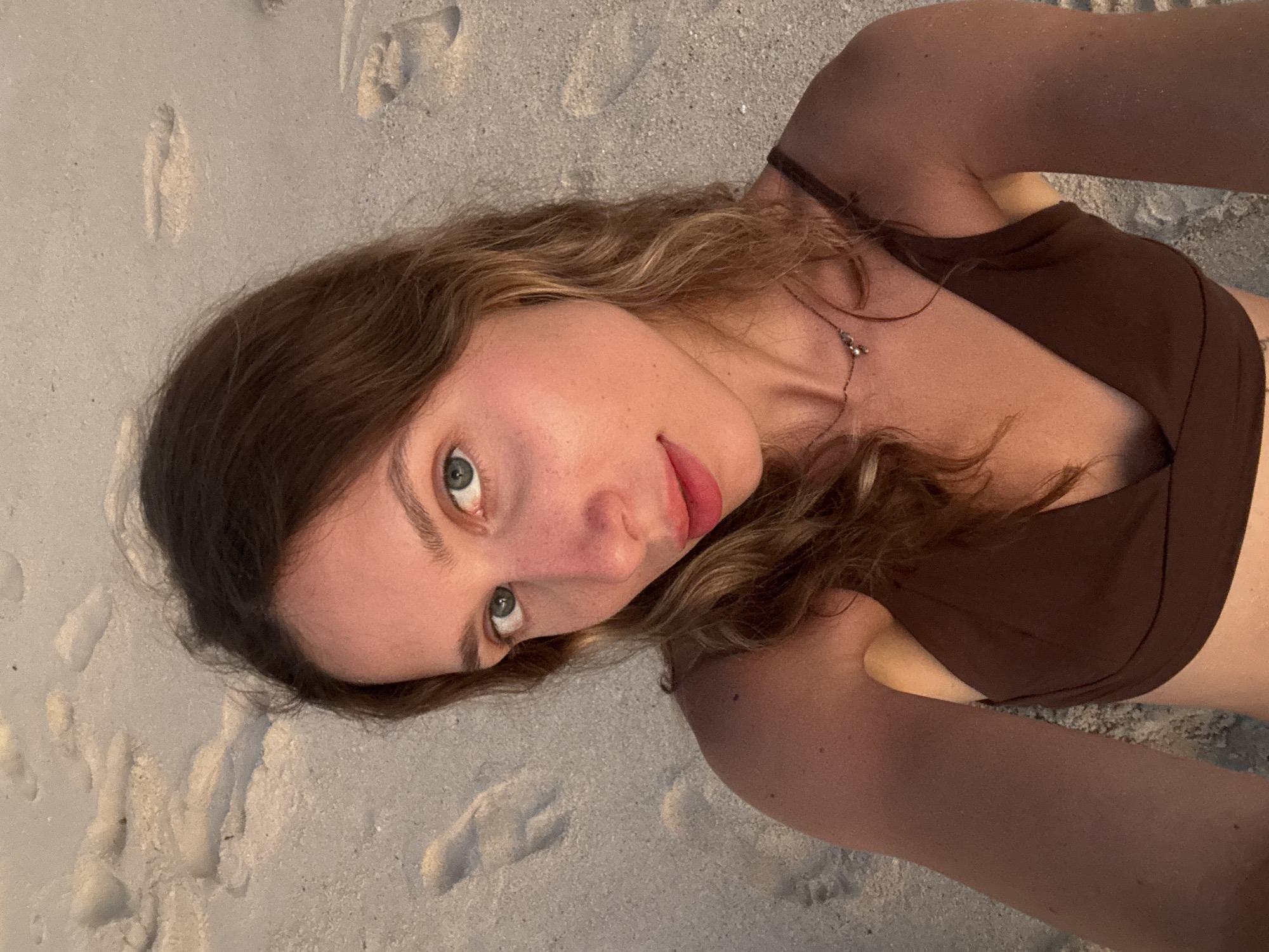 The image shows a close-up, high-angle selfie of a young woman with blonde hair and blue eyes, taken on a sandy beach. She is wearing a brown bikini top and a delicate necklace. Her face is turned slightly to her left, and she is looking directly at the camera. The background is composed of sand with visible footprints. The lighting suggests it is either sunrise or sunset, casting a warm glow on the scene. The woman's skin has a tan appearance, and there are a few freckles visible on her nose and cheeks. The overall setting is a beach in Kertasari, Indonesia.